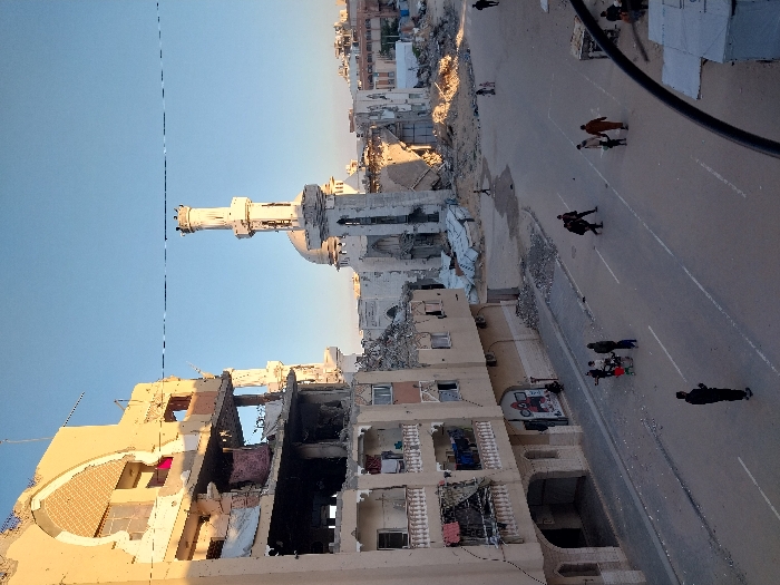 This image displays a street scene in Khan Yunis, Palestinian Territory, characterized by extensive structural damage. On the left, a multi-story building stands heavily damaged, with its upper floors showing collapsed sections, exposed interiors, and visible rebar. Laundry items are hanging from balconies on its lower, less damaged floors. In the mid-ground, a mosque features a tall, slender minaret that remains largely intact, while the main mosque building behind it and its dome exhibit significant damage and partial collapse, surrounded by piles of rubble. Further in the background and to the right, additional structures are reduced to debris.
The asphalt street, marked with two faded white lines, is covered with scattered dust and small pieces of debris. Several individuals are present on the street: a person in dark clothing stands near the left foreground, facing away from the viewer. A group consisting of approximately four adults and two children, one in a stroller or cart, walks towards the left side of the street. Further right, two individuals in dark clothing are crossing the street, and a person accompanies a child along the right side. Large piles of concrete rubble are visible adjacent to the damaged buildings and in the background areas. The sky is clear and blue. A thin, dark line, possibly a utility cable, spans horizontally across the upper portion of the frame.