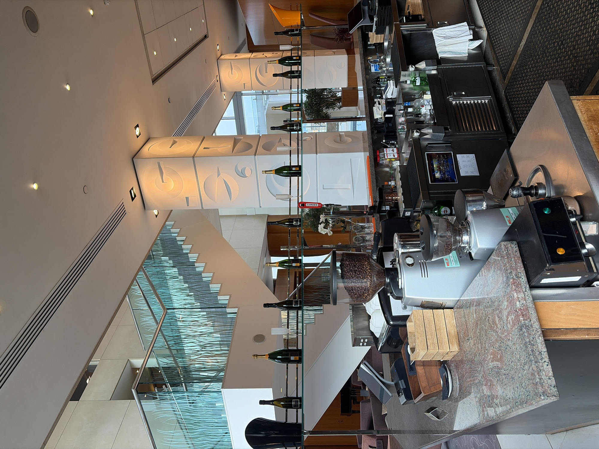 The image depicts the interior of a modern hotel lobby bar, specifically identified as being within the Montcalm Royal London House on Finsbury Square in the City of London, United Kingdom. No individuals are present in the scene.
In the foreground, a bar counter with a mottled pink and grey granite surface transitions to a stainless steel workspace. This area is equipped with professional coffee-making machinery, including a large grinder containing visible coffee beans in its hopper, a smaller grinder, and an espresso machine with a portafilter. A point-of-sale system and several wooden storage boxes are on the granite counter. Behind the main counter, a stainless steel section features sinks, various spirit bottles (including Bacardi), and a white towel hanging from an under-counter refrigeration unit. A Camden beer tap handle is also visible.
Above the bar, a clear glass shelf spans horizontally, displaying multiple dark green and black champagne bottles, several empty champagne flutes, and a black decorative vase. A small floral arrangement with white orchids is also present on this shelf.
The background features a grand white staircase with glass balustrades etched with a repetitive, wavy pattern. Several large, square architectural pillars, internally lit, are clad in white material embossed with stylized, abstract 'G' shapes or spiral motifs. Beyond these pillars, large windows suggest an exterior view, and a potted plant is visible. To the left, partially obscured by the bar, a lounge area with dark upholstered seating and a black grand piano is situated beneath the ascending staircase. The ceiling is white with recessed spotlights and linear ventilation grilles. The floor behind the bar consists of dark, textured mats.