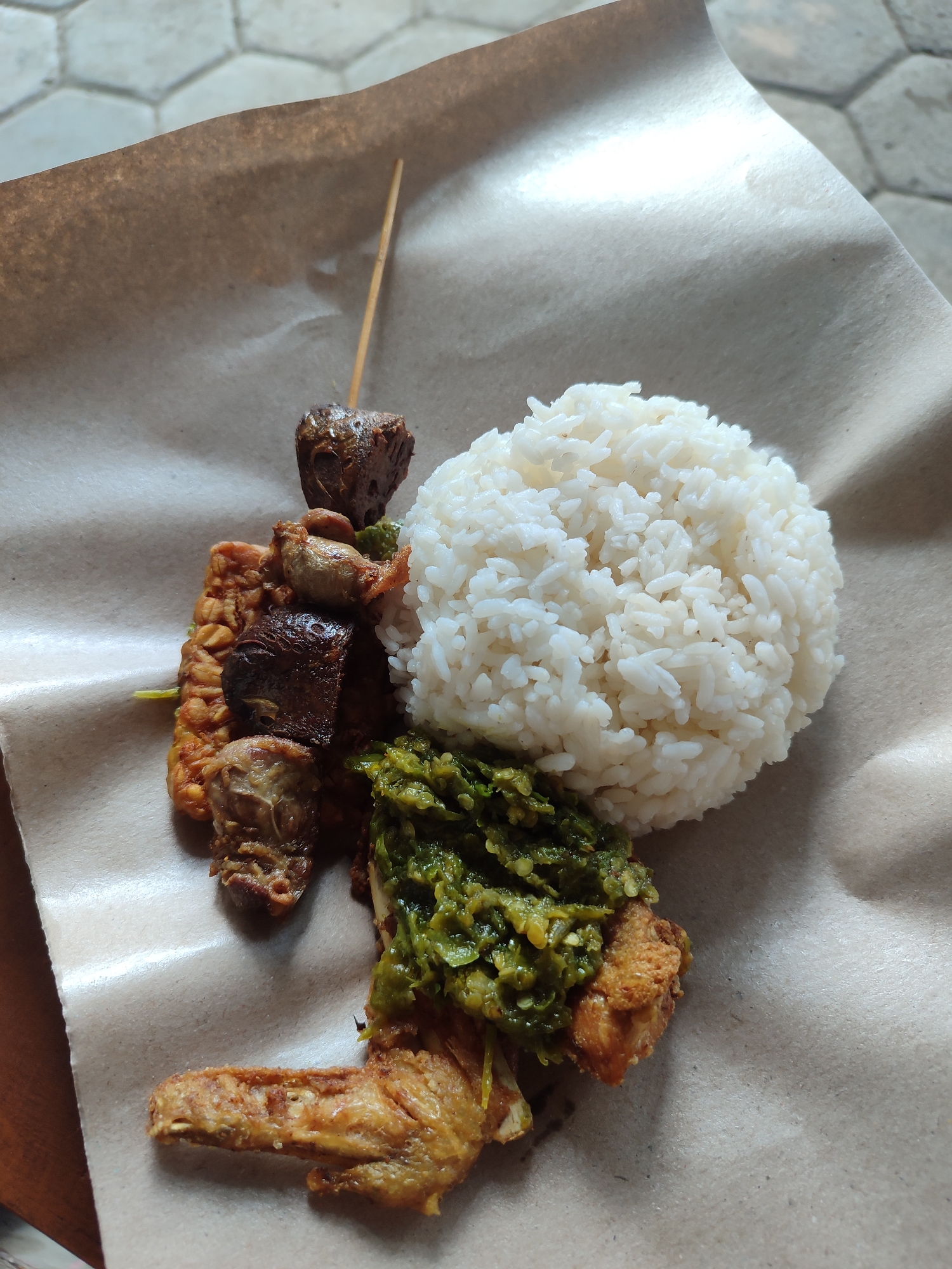 A close-up, overhead shot shows a meal served on brown paper. The meal consists of a mound of white rice, a piece of fried chicken covered in green chili sauce, and skewers of meat and tempeh. The food is arranged on a surface with a hexagonal tile pattern visible in the background. The scene is likely an outdoor setting, possibly a food stall in Kagok, Indonesia.