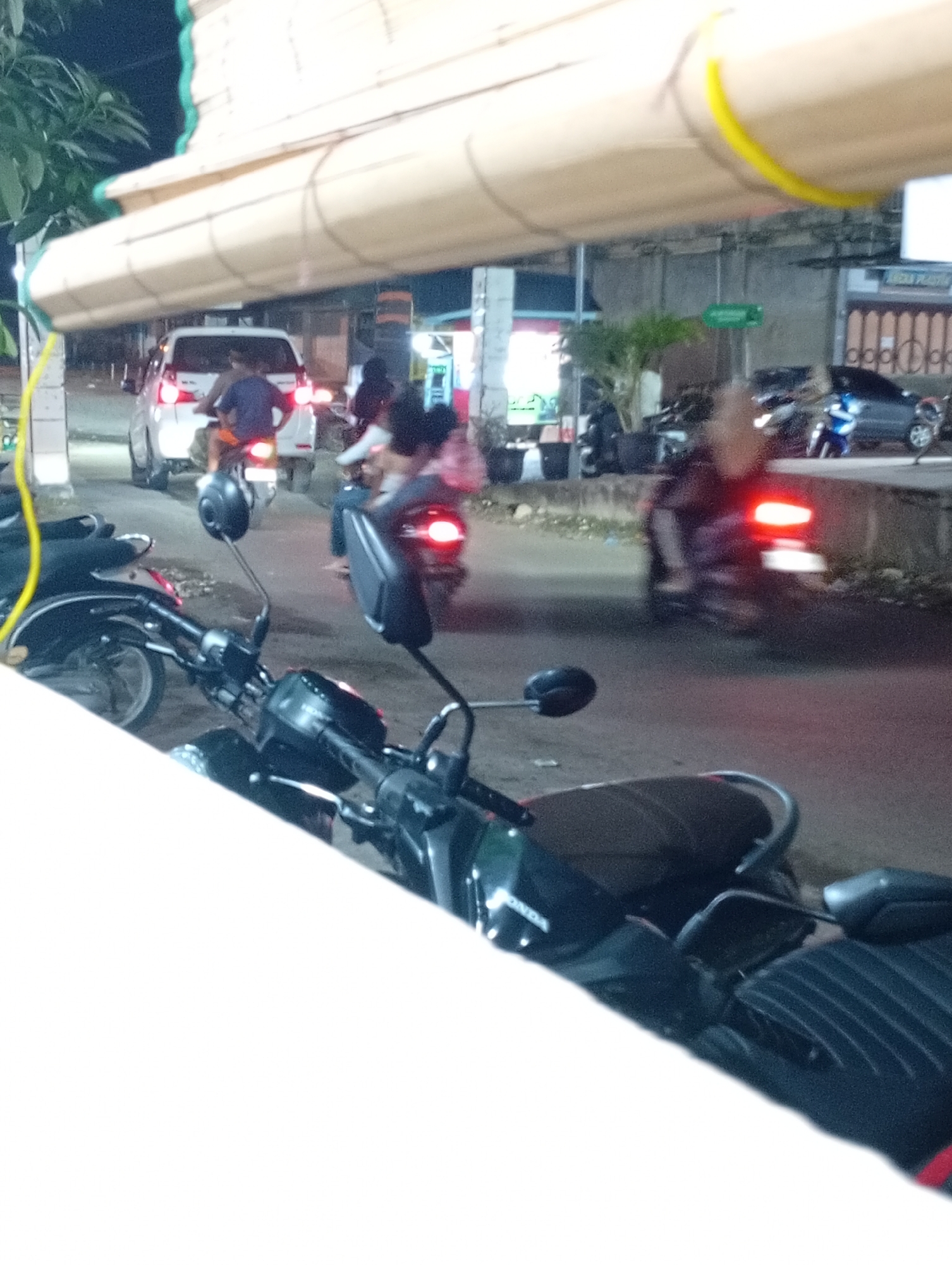 The image depicts a bustling nighttime street scene in Lhokseumawe, Indonesia, illuminated by artificial lights and vehicle head/taillights.
In the immediate foreground, the handlebars, rearview mirrors, and part of the seat of a black scooter are prominently featured, partially obscured by a bright white, out-of-focus surface in the lower left. The partial text "ONDA" is visible on the scooter's body, likely indicating a Honda brand. Another black scooter is partially visible to the left.
The midground reveals a street with several vehicles in motion or stopped. A white multi-purpose vehicle (MPV) is seen from the rear, its red taillights illuminated. Directly behind it, a person is riding a motorcycle, also with its taillight on. Other blurred figures on motorcycles, some appearing to carry passengers, suggest ongoing traffic or slow movement. To the right, a dark blue car is parked beside the road.
The background shows building facades lining the street, lit by various artificial sources. A large, light-colored roll-up awning or canopy stretches across the upper part of the frame, with a bright yellow cable or conduit running along its edge. Green foliage is visible on the far left. A very blurry sign in the distance appears to contain the text "ADDA PLAY".
The time of day is clearly night, evidenced by the darkness and the reliance on artificial lighting from vehicles and buildings. The scene conveys a sense of urban activity and motion.