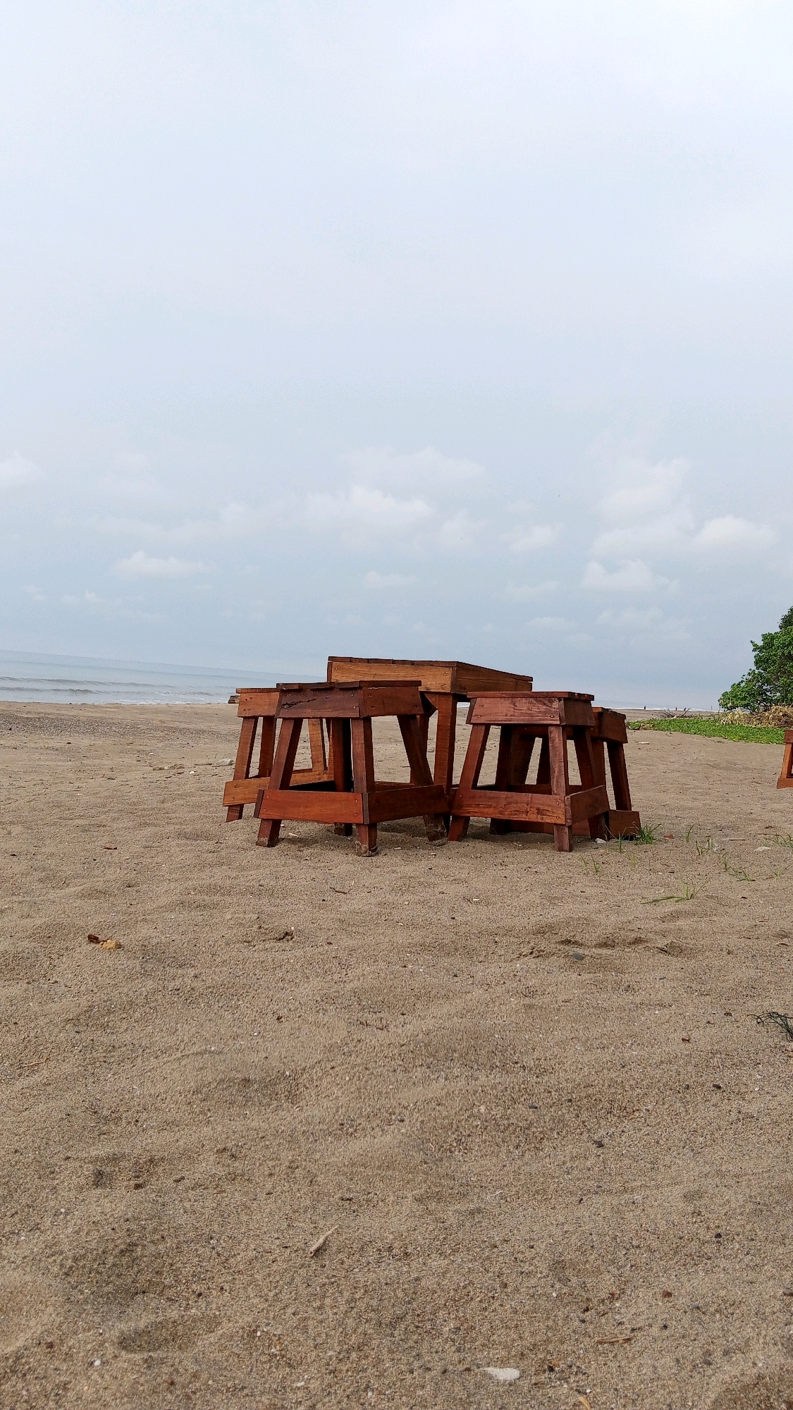 The image depicts an outdoor scene on a sandy beach in Kruengjuli Timu, Indonesia, under an overcast sky. In the foreground, a cluster of three wooden benches or tables are arranged on the sand. The furniture is dark brown and appears to be made of solid wood, with a simple, sturdy design. The benches are positioned close together, suggesting they might be intended for seating or as a surface.
The beach itself is composed of light brown sand with some scattered debris and small patches of vegetation. The ocean is visible in the background, with gentle waves rolling towards the shore. The water is a muted blue-gray, reflecting the cloudy sky. Further back on the right side of the image, a portion of a lush green tree is visible, adding a touch of natural color to the scene. The overall atmosphere is calm and somewhat deserted, with no people or animals present in the frame.