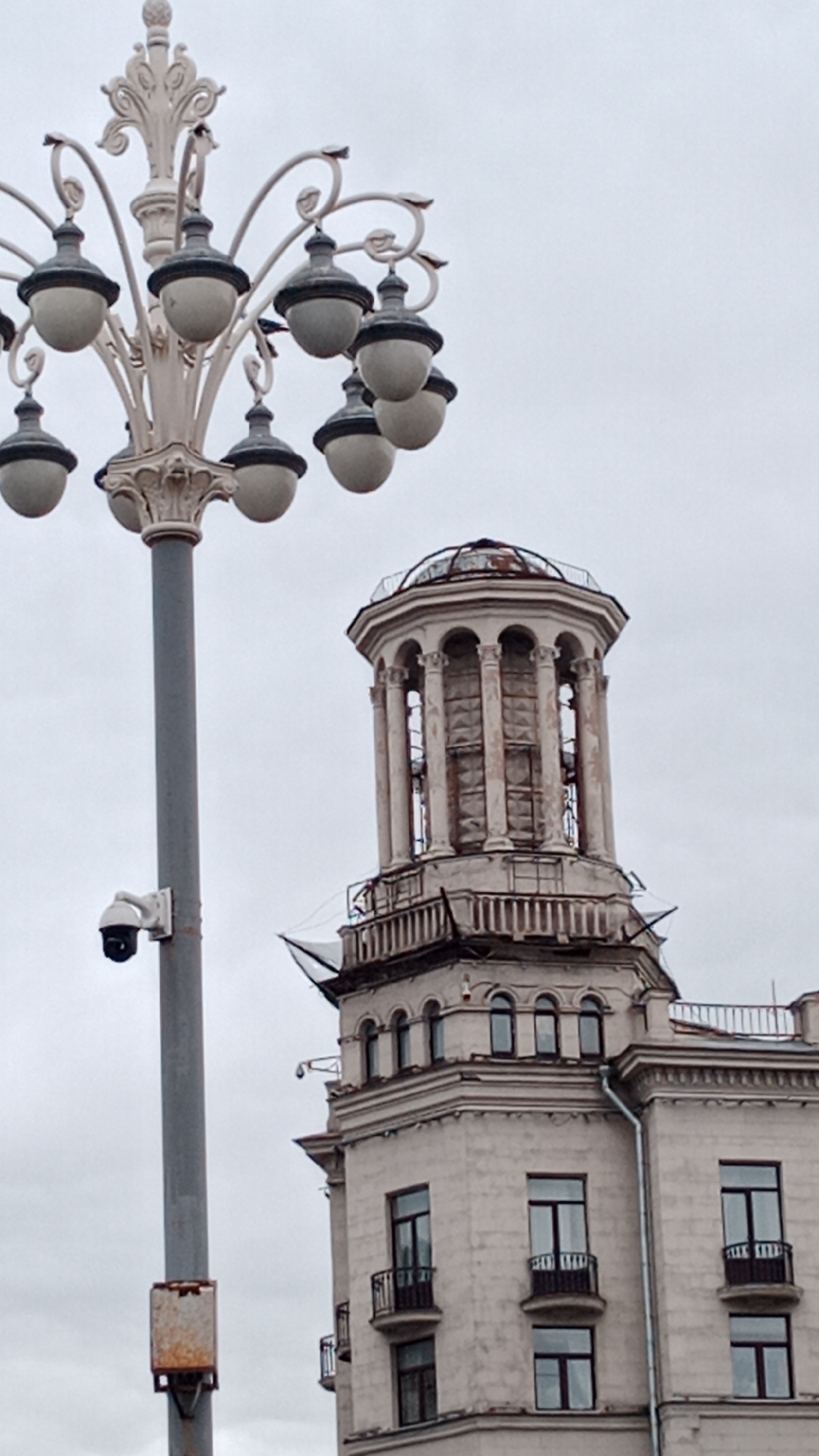 The image shows a light gray building with a tower-like structure on top. The tower has columns and a dome-shaped roof. Some scaffolding or netting is visible around the tower. In the foreground, there's an ornate lamppost with multiple globe-shaped lights and a security camera attached to it. The sky is overcast and gray. The architecture appears to be old and possibly in need of renovation. The city is not mentioned in the image. The state is not mentioned in the image.