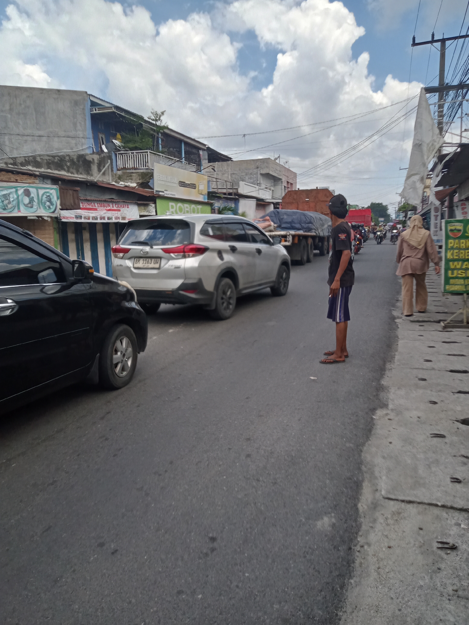 The image depicts a street scene in the city of Sumber Melati Diski, Indonesia. A black car is parked on the left side of the road, and a silver SUV is in front of it. A young man wearing a black t-shirt and blue shorts is standing on the right side of the road, facing away from the camera. Further down the road, there are several other vehicles, including trucks and motorcycles, and a woman in a beige hijab is walking on the sidewalk. Buildings line both sides of the street, with various shops and signs visible. The sky is partly cloudy.