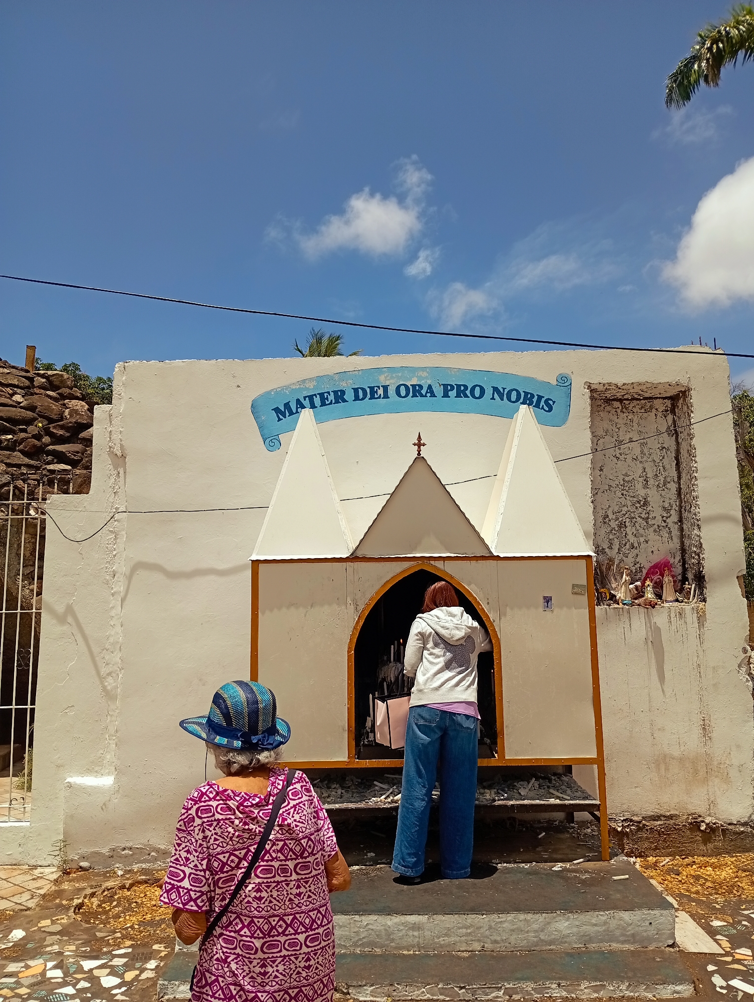 Two women stand before a small religious shrine. The woman on the left wears a patterned purple and white shirt, a blue and striped hat, and a black shoulder bag. She faces away from the shrine. The woman on the right wears a white sweatshirt, a purple shirt underneath, and blue jeans. She is placing an object inside the shrine. The shrine is a small, white building with a yellow-framed archway. Above the archway is a white, triangular roof structure and a blue banner that reads "MATER DEI ORA PRO NOBIS". To the right of the shrine is a small alcove containing several figurines. The ground in front of the shrine is paved with broken tiles. The sky is blue with a few scattered clouds. A black power line cuts across the top of the frame. The location is El Valle del Espíritu Santo, Venezuela.