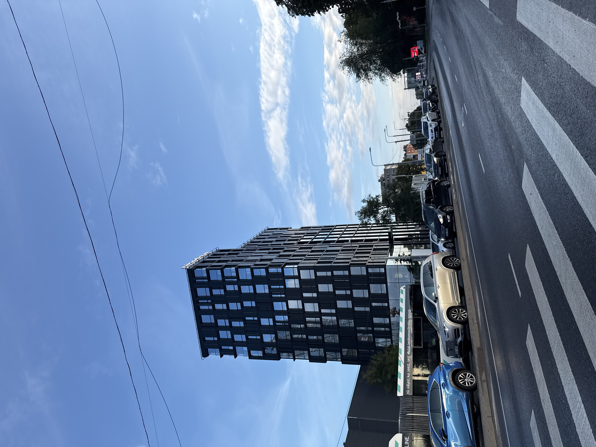 A low-angle shot captures a modern, dark-colored building with numerous windows, juxtaposed against a bright blue sky with scattered clouds. The building is tall and rectangular, with some sections protruding. In the foreground, a pedestrian crossing is visible on a grey asphalt road, with several cars parked and driving alongside it. The cars vary in color and type, including a blue SUV, a silver SUV, and several dark-colored sedans. Trees and streetlights line the road, with more buildings in the background. The overall scene suggests an urban environment in Tallinn, Estonia, likely during daylight hours.