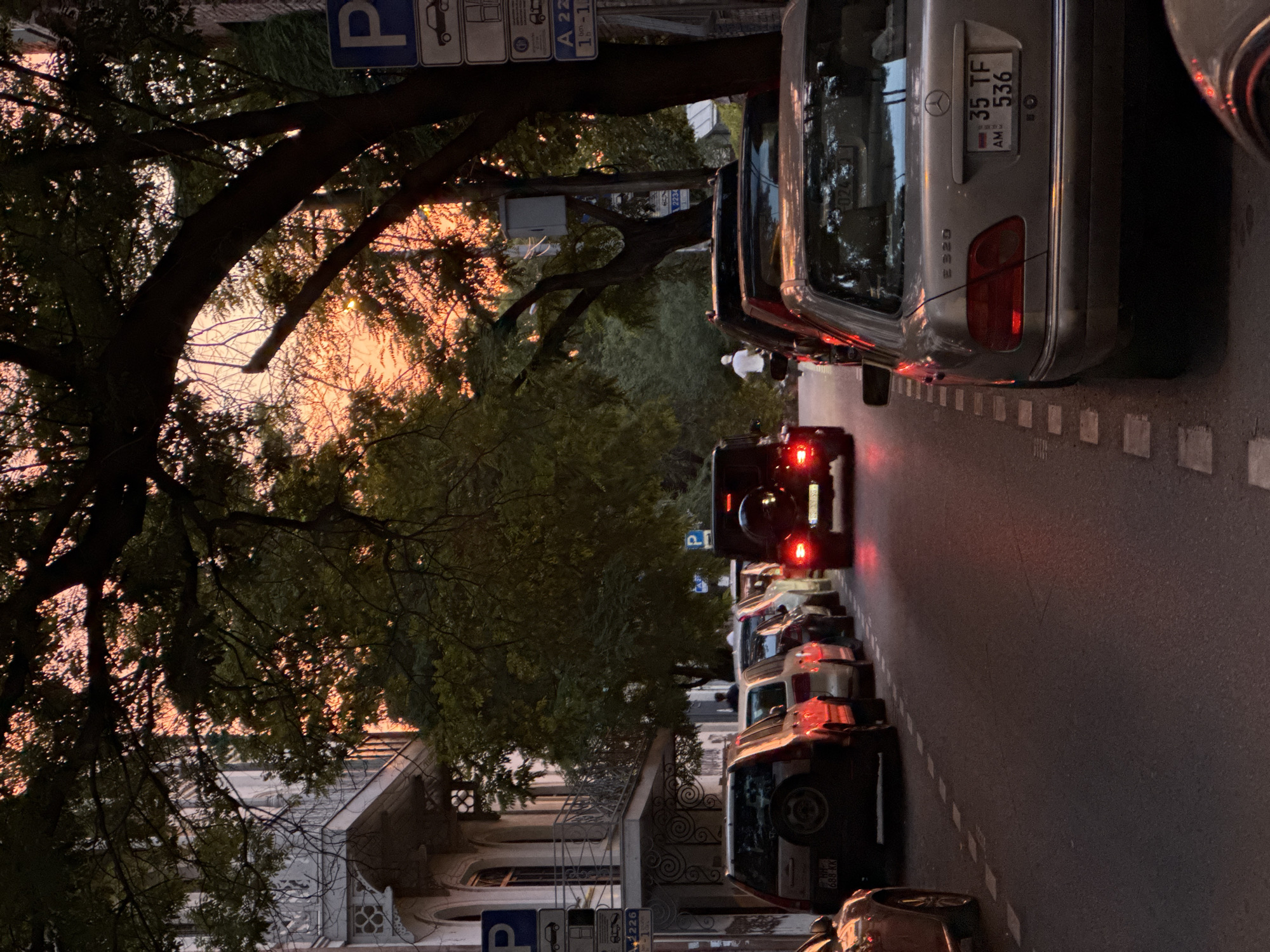 The image depicts a street scene in Tbilisi, Georgia, at dusk. Several cars are parked along the right side of the road, with a silver Mercedes-Benz E320 prominently featured in the foreground, displaying Armenian license plates "35 TF 536". A dark-colored SUV, possibly a Mercedes-Benz G-Class, is visible further down the street with its brake lights illuminated. On the left side of the road, more cars are parked, including a silver SUV with a spare tire mounted on its rear. A pedestrian in a white shirt and shorts is seen riding a bicycle in the middle of the street, heading away from the viewer. The street is lined with trees that cast shadows, and the sky is painted with the warm hues of sunset, indicating either early morning or late evening. Buildings with balconies and ornate architectural details can be seen on the left side of the street. Parking signs with the letter "P" are visible on poles along the sidewalk. The atmosphere suggests a quiet urban moment as the day transitions into night.
