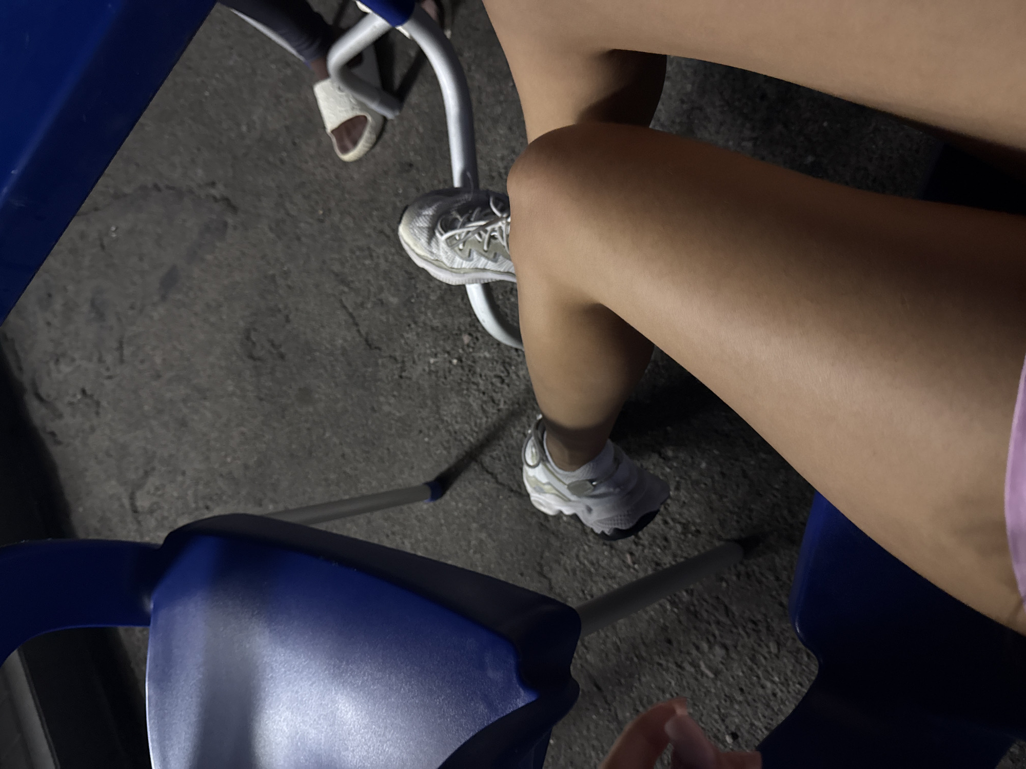 A first-person perspective shot focuses on a person's legs and feet, seated at an outdoor table. The person wears white athletic shoes and what appears to be a pink garment. The ground is asphalt, textured with small stones. In the background, another person's legs and feet are visible, wearing a dark-colored garment and white sandals. The scene is likely situated in Porumbrei, Moldova, suggested by the presence of blue plastic chairs commonly found in public spaces in the region. The lighting suggests it is either dusk or nighttime.
