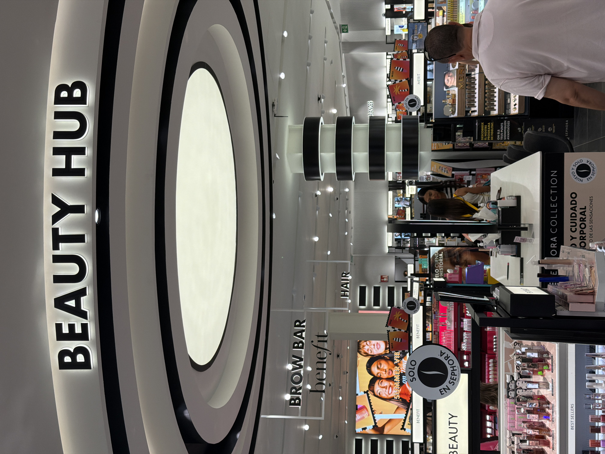 The image shows the interior of a Sephora beauty store, likely located in Barcelona, Spain, as indicated by signage in Catalan and Spanish. Above the store's entrance, a large illuminated sign reads "BEAUTY HUB". Further inside, another sign indicates a "BROW BAR" by Benefit Cosmetics. Products, primarily makeup and skincare, are displayed on shelves and tiered stands throughout the store. The scene captures the bustling retail environment with several individuals browsing or working, including a man in a light pink polo shirt facing away from the camera, and two women on the left side of the image, one of whom appears to be an employee. Promotional materials featuring models and product close-ups are visible on digital screens and displays. The store features modern interior design elements, including sleek black and white circular lighting fixtures and stacked cylindrical displays.