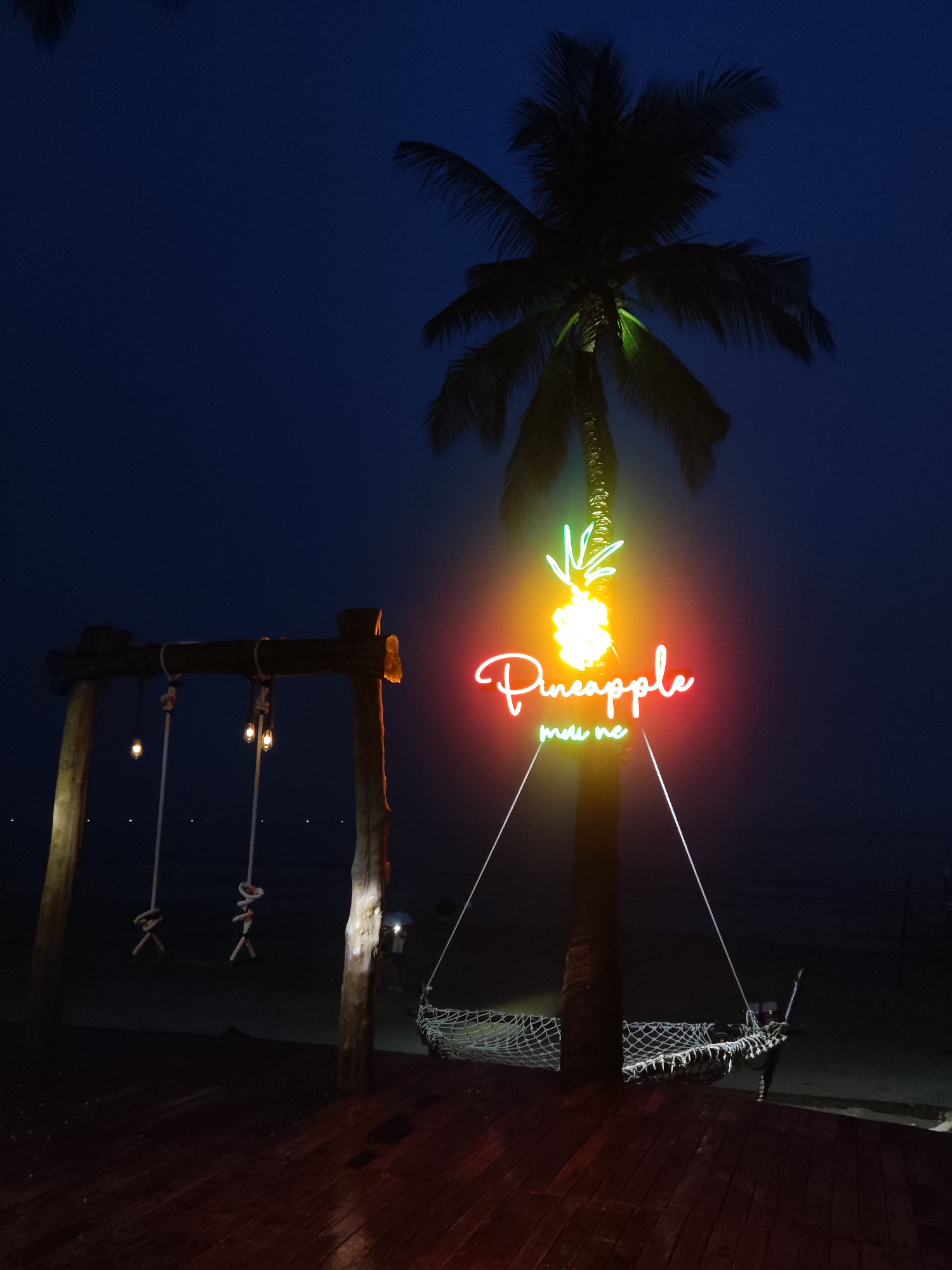 A photograph taken at night in Unknown, Unknown, shows a palm tree with a neon sign reading "Pineapple mui ne" in red, with a green neon pineapple on top. The palm tree also supports a hammock strung between it and the wooden deck. A swing set with hanging ropes is to the left of the palm tree. The background is a dark body of water and a dark sky. The overall scene is dimly lit, relying on the neon sign and the light coming from the swing set's bulbs to illuminate the area.