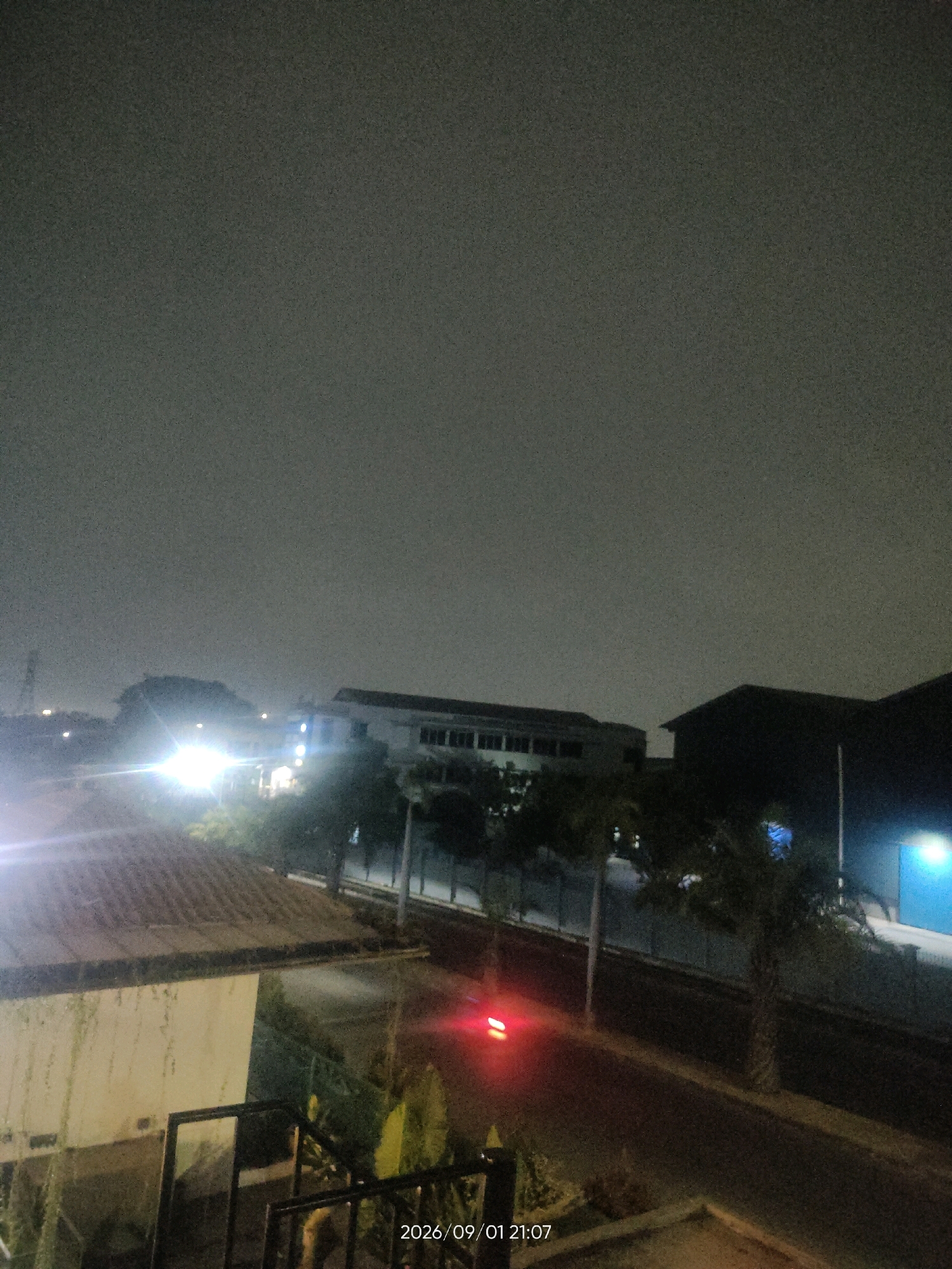 A nighttime aerial view of a street in Telagaasih, Indonesia, features a dark, overcast sky. A building with multiple windows is visible on the left, illuminated by bright lights. A street runs horizontally across the image, with a single red light, possibly from a vehicle, visible in the distance. Palm trees are scattered along the street and around the buildings. A portion of a tiled roof is in the foreground on the left. The timestamp "2026/09/01 21:07" is displayed in the lower right corner of the image.
