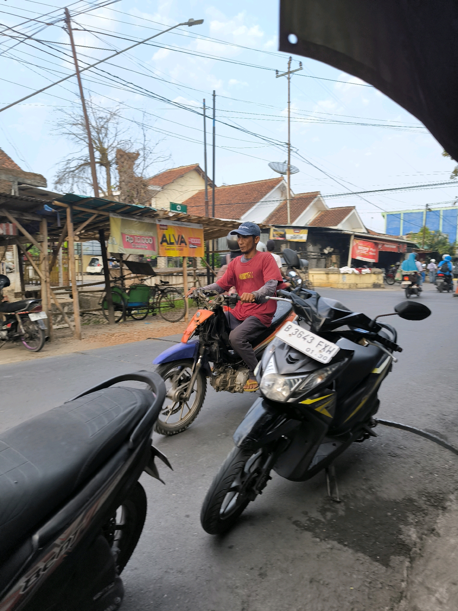 In Karanganyar, Indonesia, a man wearing a red shirt and blue cap rides an orange and blue motorcycle. He is on a street lined with buildings and stalls. In the foreground, a black motorcycle is parked. In the background, several other people are seen riding motorcycles, and there are also bicycles. The sky is visible above with scattered clouds. The overall scene depicts a busy street in a local Indonesian city.