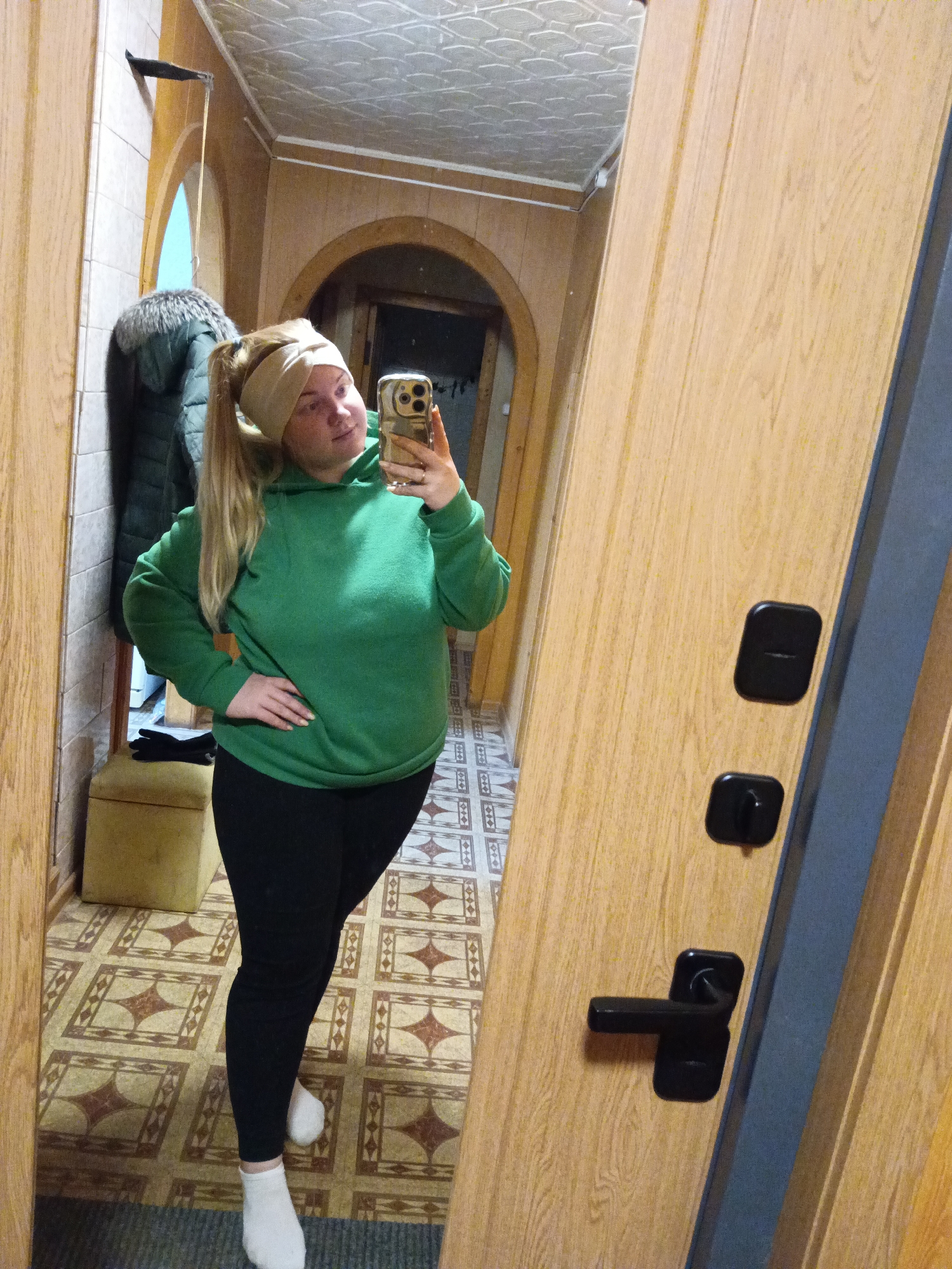 A young adult female stands in a hallway or entryway taking a selfie in a mirror. She is wearing a green hooded sweatshirt, black leggings, and white socks. Her blonde hair is pulled back in a high ponytail and she is wearing a light-colored headband. She is holding a smartphone in her right hand. The hallway has patterned linoleum flooring and light wood-paneled walls. There is a coat rack with a green jacket on the left side of the image. A small upholstered ottoman is also visible. A doorway is visible in the background. The scene is in an interior location in Lukintsy, Russia.