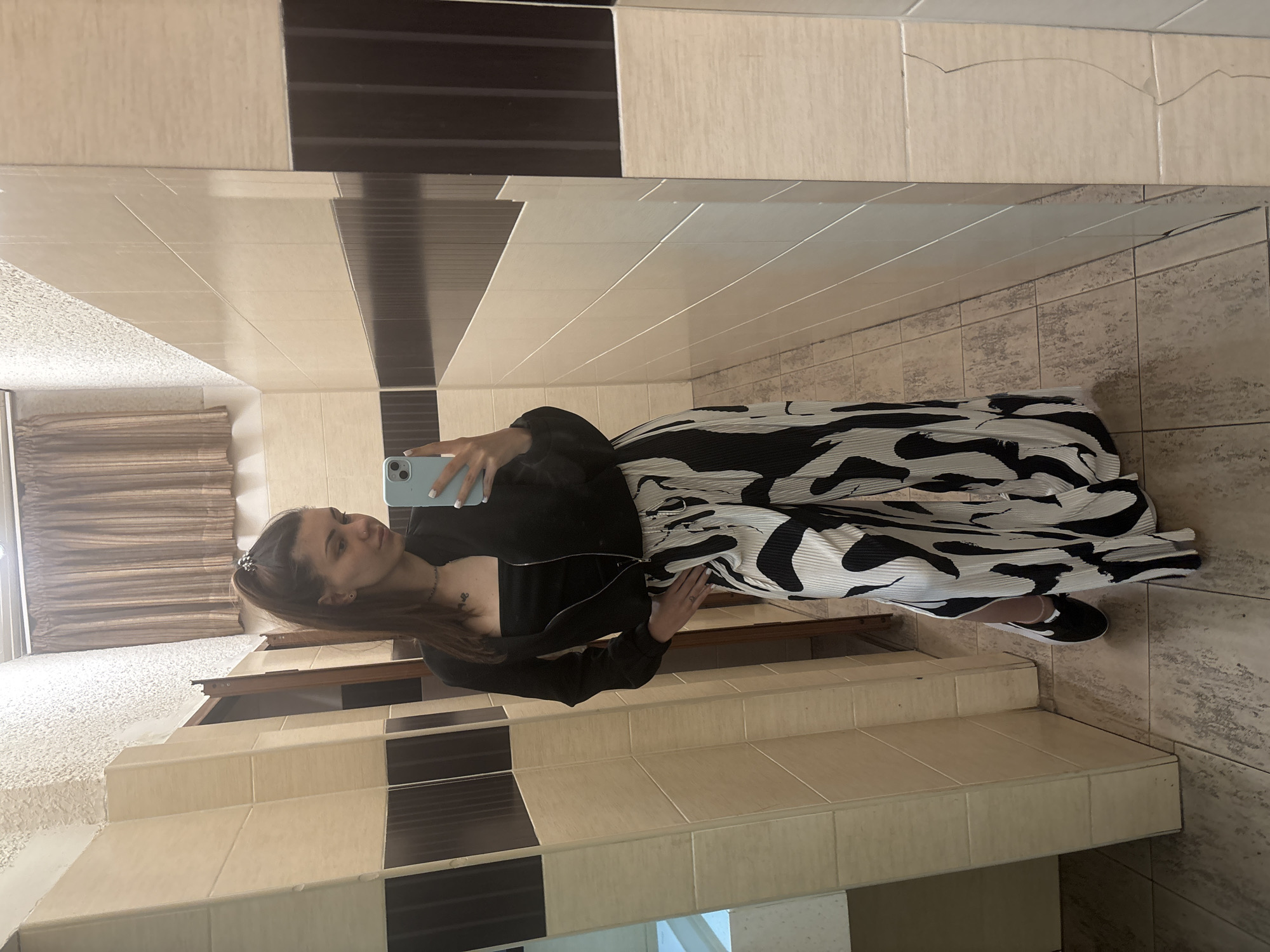 A woman is standing in a bathroom, taking a picture of herself in a mirror. She is wearing a black jacket and a black and white patterned skirt. She is holding a light blue phone. The bathroom has tiled walls with a black and white decorative stripe near the top. The floor is also tiled. The image provides no information that can identify its location in Sofia, Bulgaria.