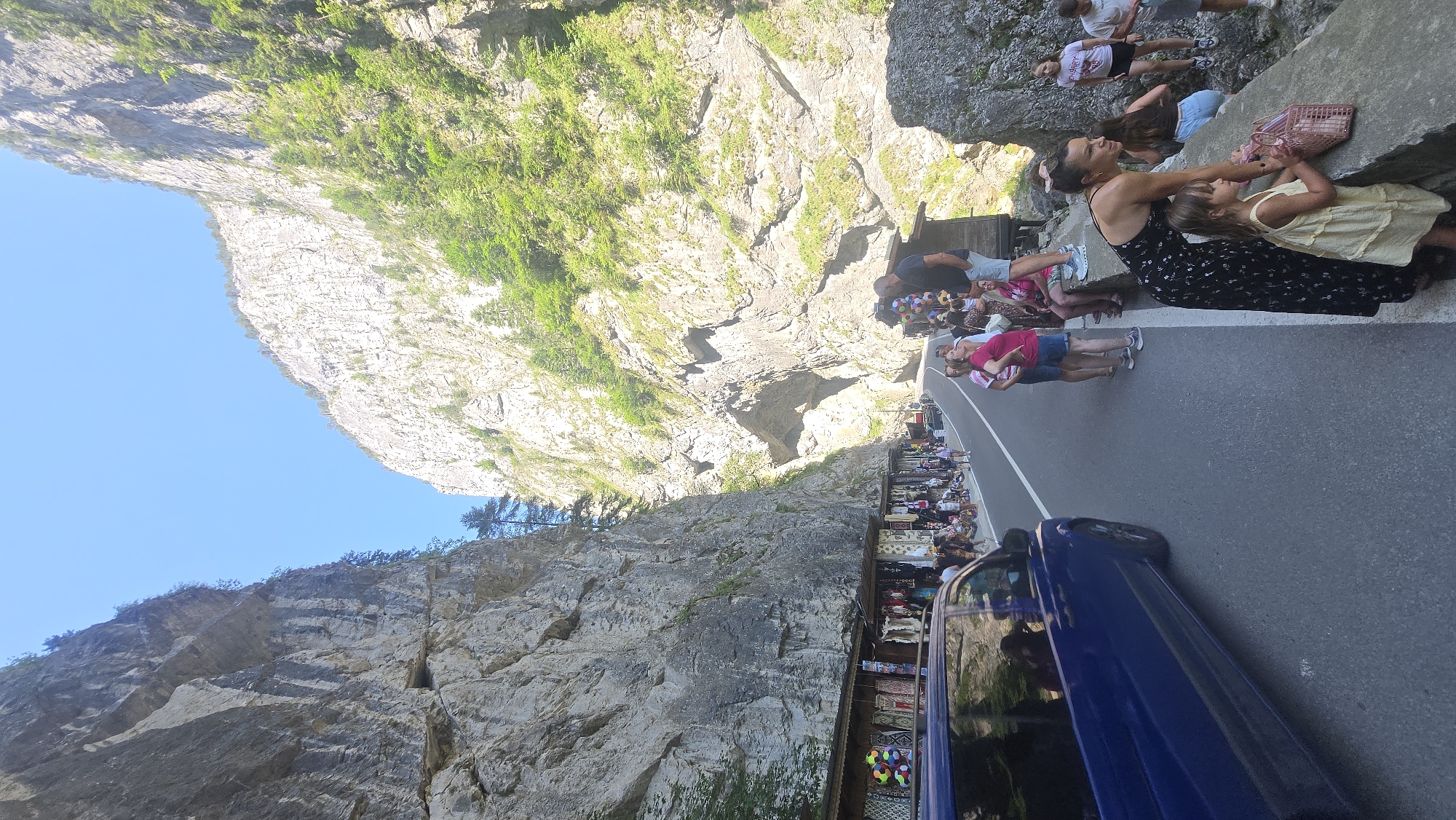 The image depicts a scene in the Bicaz Gorge, near Bicaz-Chei, Romania, on a sunny day. A paved road runs through a narrow gorge with towering, rocky cliffs on either side. On the left side of the road, several stalls are set up selling various items, with people browsing and shopping. On the right side of the road, a group of tourists stands along a stone wall, observing the scenery. A blue van is visible on the left, driving away from the camera. The cliffs are covered in vegetation in some areas, and the sky is clear blue. The overall atmosphere appears to be that of a popular tourist destination.