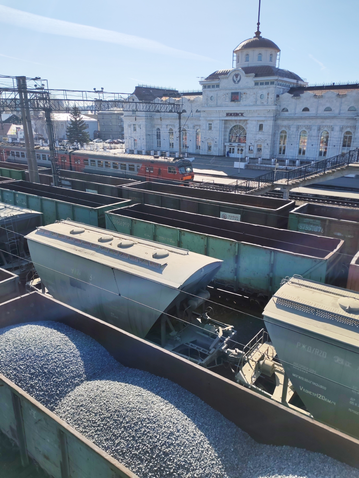 The image presents an elevated view of a bustling railway station and its surrounding tracks in Izhevsk, Russia, under a bright, clear sky. The perspective looks down upon numerous train cars in the foreground and midground, extending towards the main station building in the background.
In the immediate foreground, an open-top freight car is prominently featured, filled with light grey crushed stone or gravel, forming two distinct mounds. Behind it, a light-colored covered hopper car, possibly for bulk materials, shows several hatches on its roof and the partial number "912 3". Further back, a line of green open-top gondola cars stretches across multiple tracks, with some displaying numbers such as "55956999" and "57557876", and one bearing a "modum" logo. Another hopper car is partially visible with detailed Russian text, including "РЖД/RZD", "ПОСТРОЕН 12194" (Built 1994, likely a typo in OCR for 2014 or similar), "МОГИЛЕВВАГОНЗАВОД" (Mogilev Wagon Plant), and "V КОНСТР120км/ч" (Construction speed 120 km/h).
In the midground, a red and grey passenger train is parked on a track adjacent to the station platform. The main subject in the background is the grand Izhevsk railway station building, a white or light-colored stone structure featuring a prominent dome with a spire and a digital clock tower displaying "15 26". The name "ИЖЕВСК" is clearly visible above the main entrance. A solitary person in a blue uniform or jacket is seen near the station entrance on the platform. Overhead electrical lines and railway infrastructure, including signal gantries, crisscross the scene. Beyond the station, other urban buildings and scattered trees are visible, with a notable evergreen tree near some residential structures.
The bright sunlight and clear blue sky indicate a sunny afternoon. The scene captures the functional activity of a railway hub, with both freight and passenger services, under calm weather conditions.