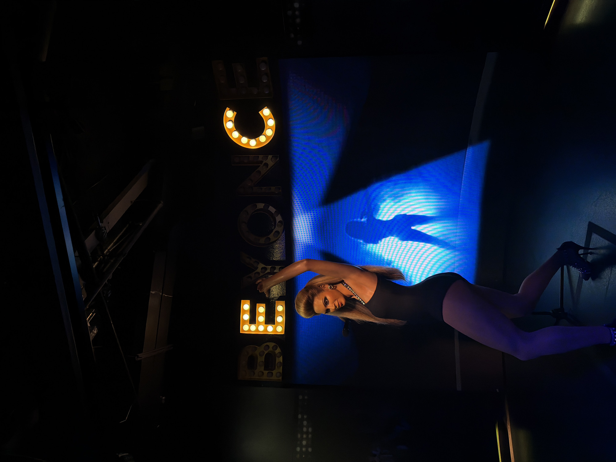 The image shows a wax figure of a woman, appearing to be Beyoncé, standing on a stage. She is wearing a black one-piece outfit and high-heeled shoes, with her hair styled in a bouffant. Behind her, a large blue screen displays a silhouette of a dancer and the word "BEYONCE" in illuminated marquee letters. The lighting is dramatic, with spotlights illuminating the figure and the background. The scene is set in a dimly lit indoor venue, likely a museum or entertainment attraction. There is no indication of the specific location in Bangkok, Thailand.