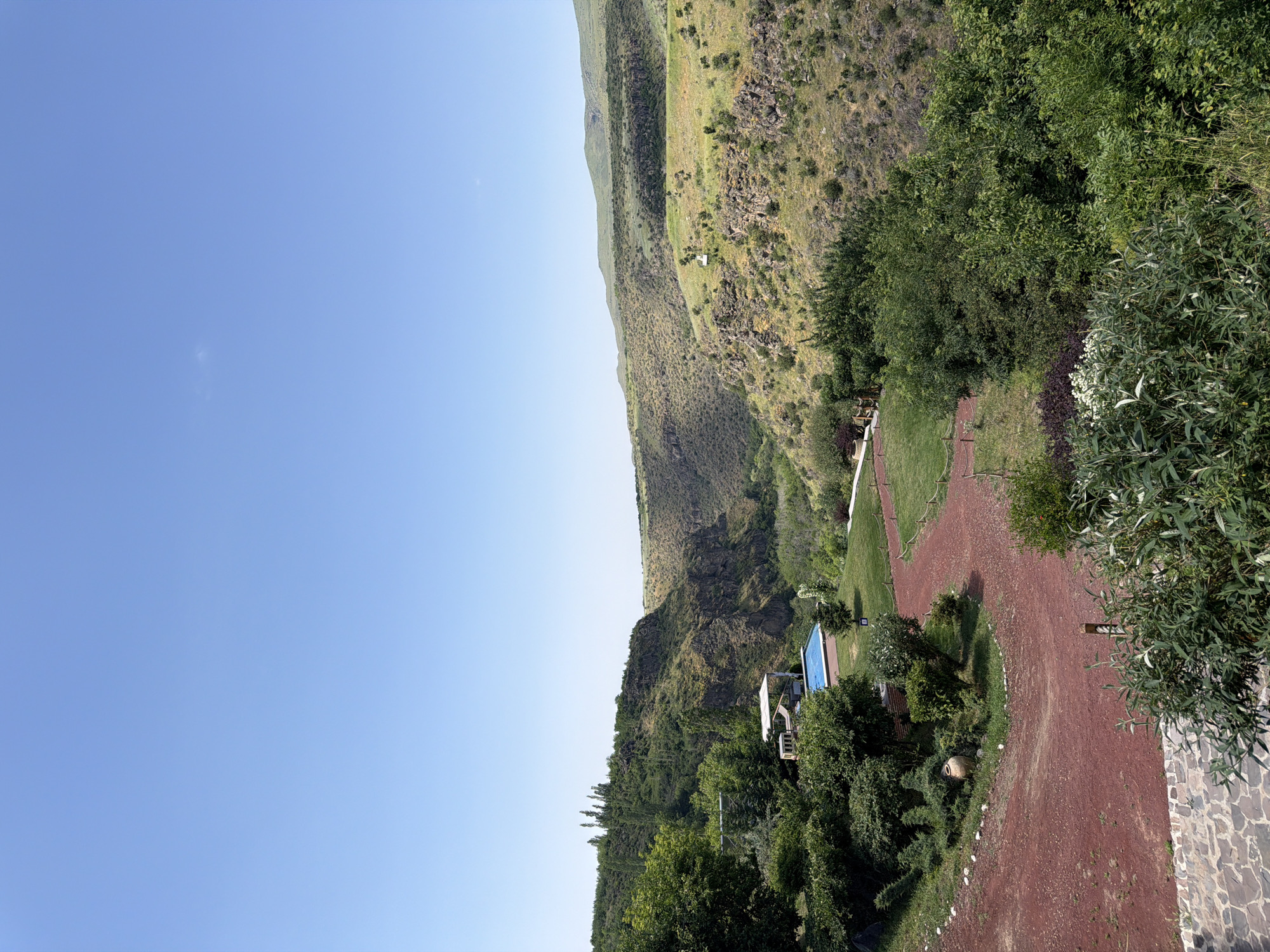 This image captures a scenic view in Byurakan, Armenia, featuring a natural landscape with man-made elements. The foreground shows a winding path made of reddish gravel, bordered by lush green bushes and trees. In the mid-ground, a swimming pool with a blue cover is visible, adjacent to a white structure with a canopy, surrounded by manicured green lawns and trees. Further in the distance, a deep canyon carves through the terrain, with steep, rocky slopes covered in sparse vegetation. Rolling hills with a mix of green and brown hues form the backdrop, under a clear, bright blue sky. The overall scene suggests a tranquil setting, possibly a resort or a private residence situated amidst the natural beauty of the Armenian countryside.