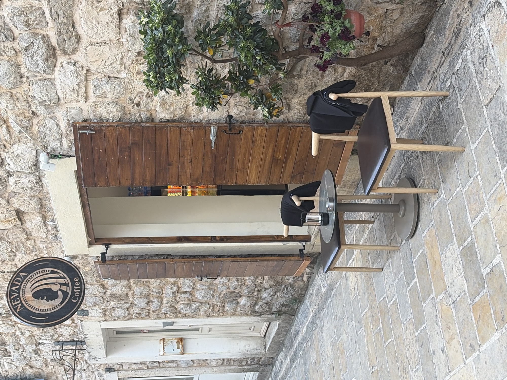The image shows the exterior of Nevada Coffee in Budva, Montenegro. The building has a stone facade with a wooden door and a small window. A black jacket and a drum are placed on the chair. A small table is next to the chair. The street is paved with light-colored stones. A Nevada Coffee sign is on the side. A small tree grows above the door.