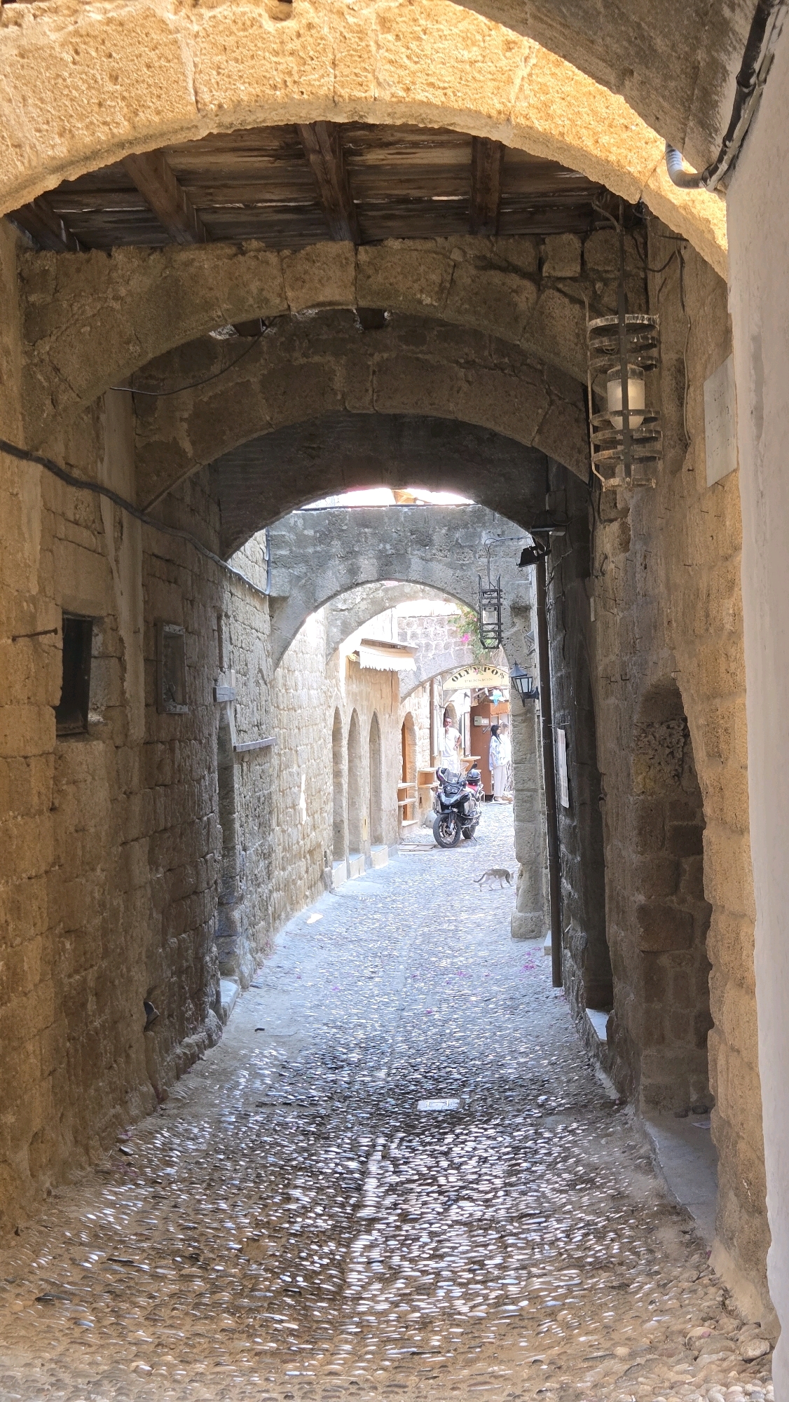 This is a sun-drenched view down a narrow, arched cobblestone alleyway in the Municipal Unit of Rhodes, Greece. The stone walls on either side are made of rough-hewn, tan-colored blocks. The alley is covered by multiple stone arches, one of which forms a substantial entryway with a wooden ceiling above. A rustic metal lantern hangs from the upper archway. The cobblestone path is uneven and glints with reflected sunlight, dotted with small pink petals. In the distance, a black motorcycle is parked on the right side of the alley. Two figures, one in white clothing, are visible further down the alley. A small cat is walking across the cobblestones near the motorcycle. The scene evokes a sense of ancient history and Mediterranean charm.