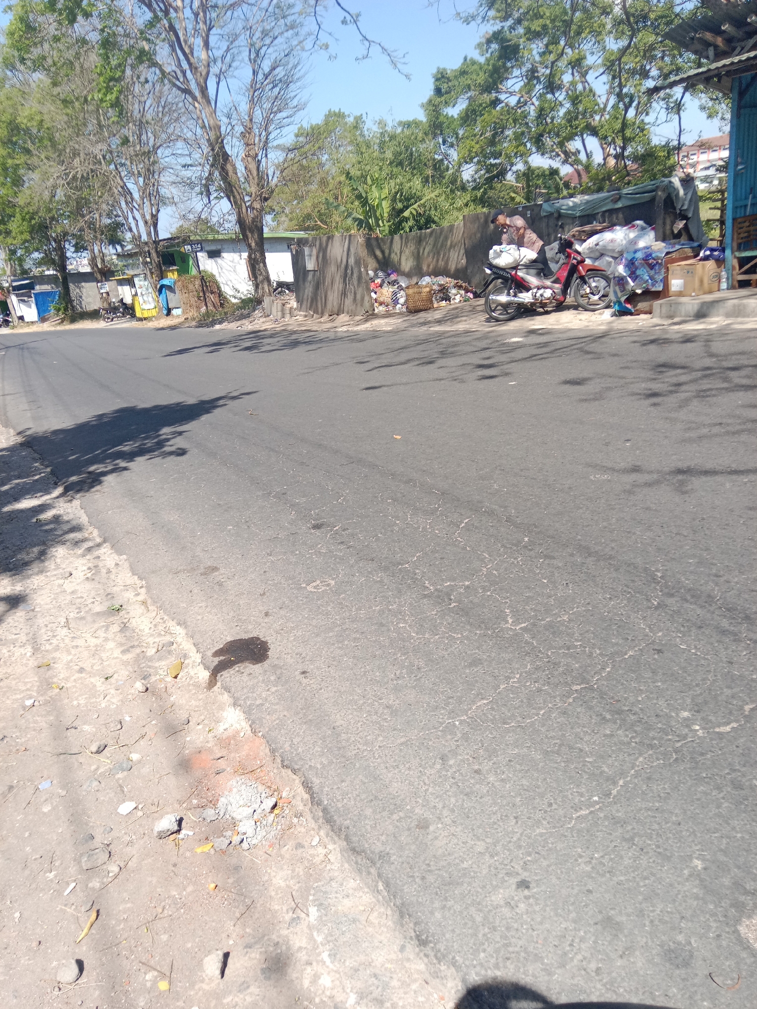 A medium shot captures a sunny day in the city of Dangkel, Indonesia. The image depicts a dusty roadside with a paved asphalt road dominating the foreground. To the left of the road, a sandy, unkempt shoulder is visible, littered with small rocks and dry leaves. A dark, oil-like stain mars the asphalt in the foreground, casting a shadow that resembles an abstract shape.
In the background, a line of trees with sparse foliage stands between the road and a row of buildings. One building, possibly a shop, is painted in various bright colors, including blue and yellow. A person, wearing a blue shirt and dark pants, stands near this building.
Further down the road, a red and silver motorcycle is parked. A person in a light-colored shirt and dark pants is leaning over the motorcycle, seemingly adjusting something or attending to it. Several large plastic bags filled with what appears to be trash are piled next to the motorcycle and a low concrete wall. A cardboard box is also present. The background shows more trees and buildings, with a slightly blurred building in the distance. The sky is clear and blue. The scene conveys a sense of everyday life on a street in a tropical urban environment.