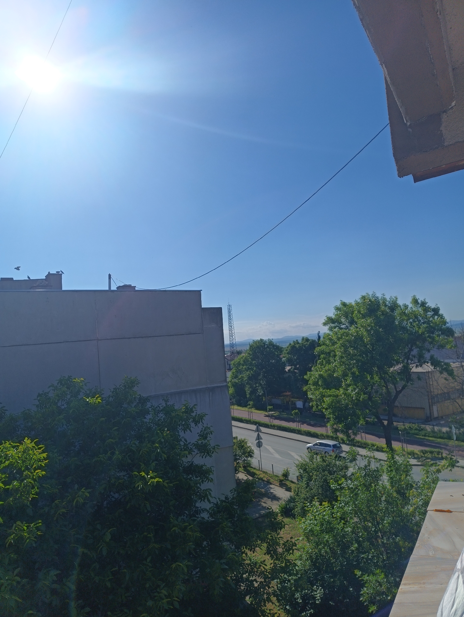The image depicts a sunlit urban scene with a clear blue sky dominating the upper portion. In the foreground, the corner of a concrete building juts into the frame from the top right. A thin dark wire stretches diagonally across the sky from the upper left to the lower right. To the left, a large, gray, flat-roofed building with horizontal paneling occupies a significant part of the mid-ground. Several birds are visible on the roof of this building. In front of the gray building and extending towards the bottom of the frame are lush green trees.
In the background, a street with a silver car driving on it is visible, bordered by a sidewalk and more trees. Beyond the street, there are industrial-looking buildings and a tall, lattice-like structure resembling a telecommunications tower. Distant hills are visible on the horizon under the bright sun. The scene appears to be a view from an elevated position, possibly a balcony or window, overlooking a residential and commercial area. The lighting suggests it is a bright, clear day. The location is identified as Karnobat, Bulgaria.