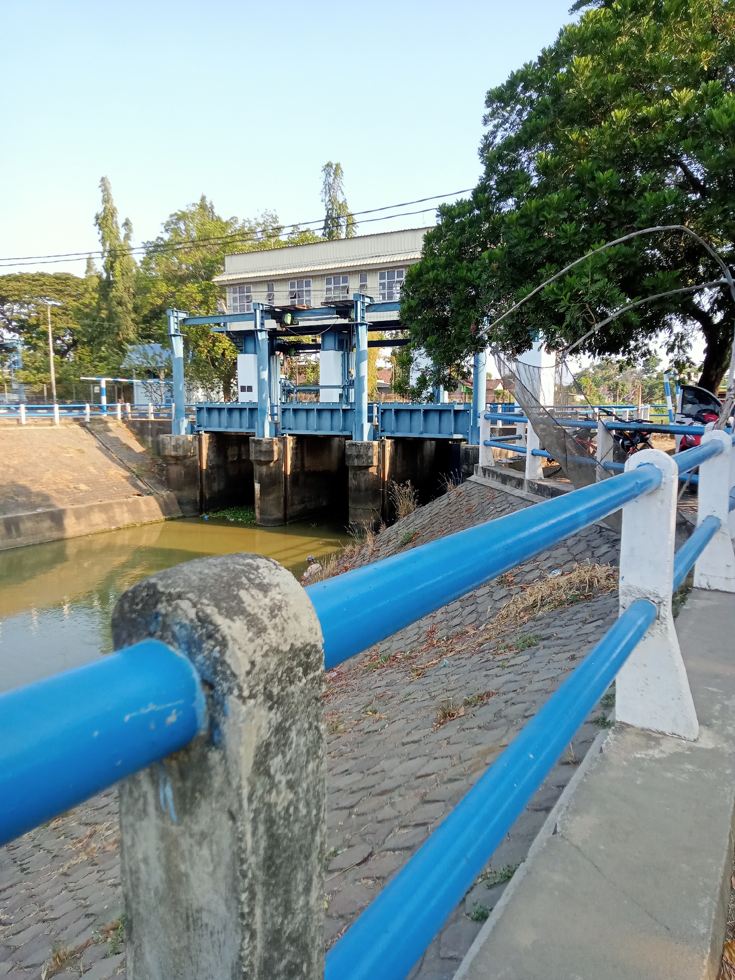 The image shows a water gate structure in Majan, Indonesia. The gate, painted blue, is situated across a canal filled with murky green water, with some aquatic plants visible. The structure appears to be made of concrete and metal. In the foreground, a bright blue metal railing with white concrete posts runs along a paved walkway. Beyond the water gate, there is a building with a light-colored roof and windows, surrounded by lush green trees. The sky is clear and blue. In the background, other trees and some parked motorcycles are visible. The overall scene depicts an infrastructure facility in a natural environment.
