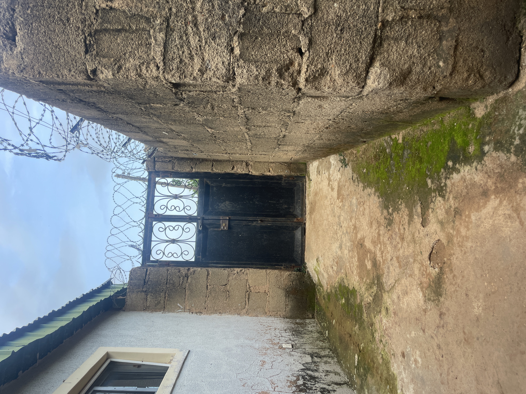 This image depicts an outdoor scene in Bukuru, Nigeria. In the center of the frame is a black metal gate with an ornate design, situated between two walls. The wall on the left is a lighter color, possibly stucco or painted concrete, with some cracking and staining visible. It has a window with a white frame. The wall on the right is constructed from large, rough-textured concrete blocks. Both walls are topped with razor wire fencing, suggesting a security measure. The ground is a mixture of concrete and dirt, with patches of moss and some puddles, indicating damp conditions. The sky above is partly cloudy. The overall impression is of an alleyway or pathway leading to a secured entrance.