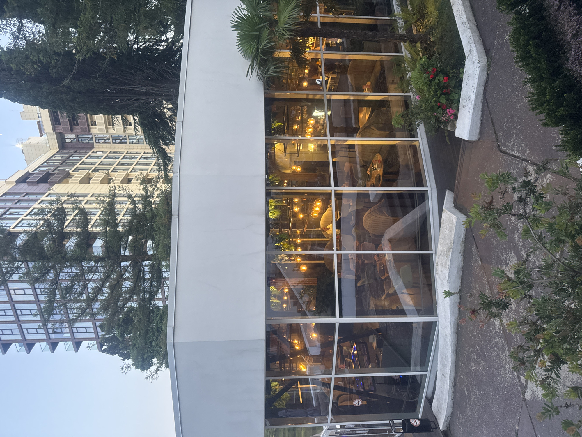 The image captures a modern building with large glass windows, likely a restaurant or cafe, in Sochi, Russia. Inside, warm lights illuminate the space, revealing glimpses of furniture such as tables and chairs, suggesting an interior dining or lounge area. The windows also reflect the surrounding environment, including trees and other buildings. A palm tree stands prominently near the building, a common sight in Sochi. The exterior of the building is white, contrasting with the glass and the greenery outside. The time of day appears to be evening or dusk, indicated by the artificial lighting within the building and the dim natural light. The weather seems calm. There are no visible text elements in the image.