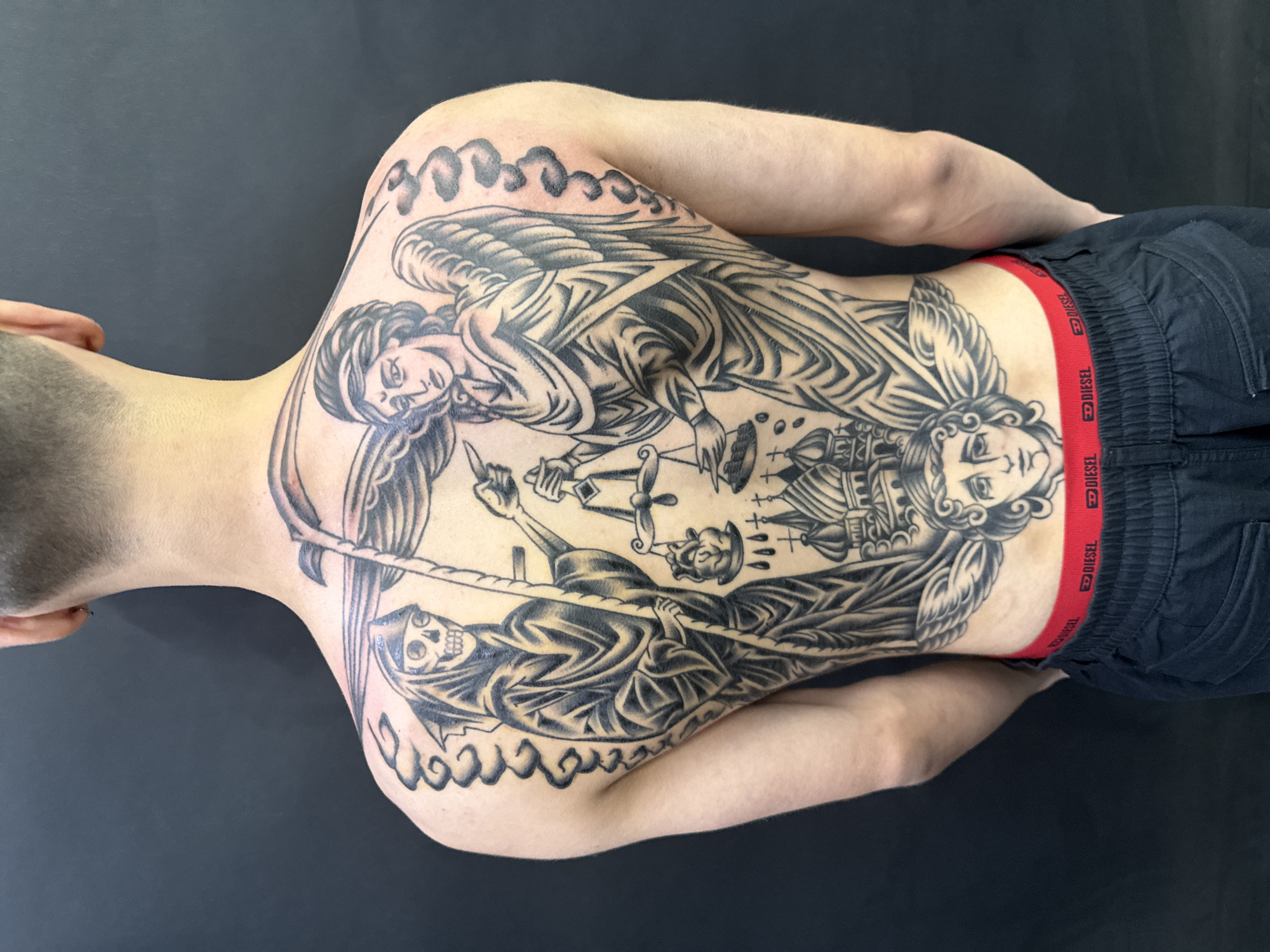 The image shows the back of a person with an elaborate black and grey tattoo covering their entire back. The tattoo depicts a Grim Reaper figure on the left, holding a scythe and a scale, with a skull for a head and a hooded robe. On the right, an angelic figure with wings and a flowing robe holds one side of the scale, which is weighing a heart. Below the figures are stylized depictions of Russian Orthodox churches with domes, and an angelic face with wings. The tattoo art style appears to be traditional or neo-traditional. The person is wearing black pants with a red waistband that has "DIESEL" branding. The background is a solid dark color, suggesting a studio or a plain backdrop. There is no discernible location in Moscow, Russia, visible in the image.