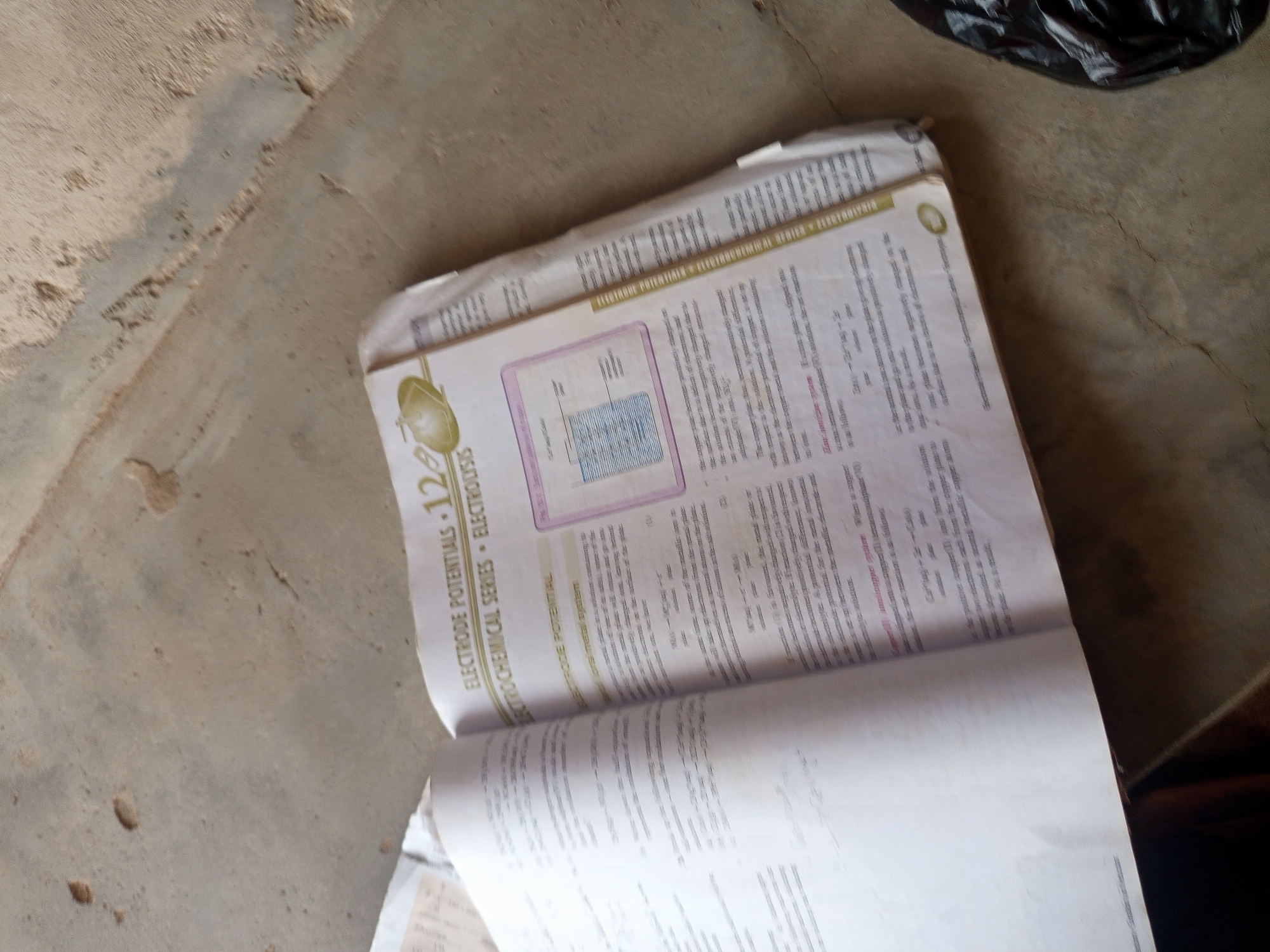An open book is placed on a concrete surface. The book is open to a page with text and an illustration of an electrochemical cell. The text discusses electrode potentials and the electrochemical series. The illustration depicts a metal plate immersed in a solution with a scale indicating electrical charge. The concrete surface appears to be outdoors, with some dirt and debris visible. The lighting suggests it is daytime.