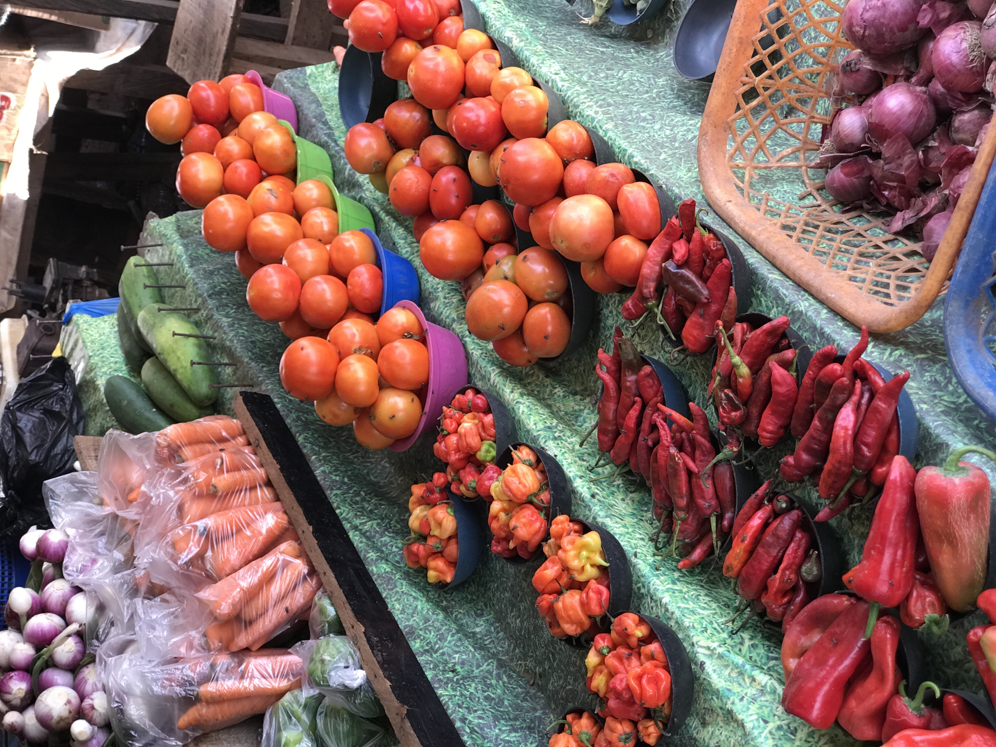 This is a photograph of a fruit and vegetable stall, likely in a market setting. The location is identified as Abuja, Nigeria.
The stall displays a variety of fresh produce, including tomatoes, red onions, carrots, cucumbers, red bell peppers, and several types of chili peppers. The tomatoes and chili peppers are arranged in piles within colorful plastic and metal bowls, while the carrots are packaged in clear plastic bags. The red onions are placed in a woven basket. The produce is set on a surface covered with a green and black patterned cloth or material. Wooden structures are visible in the background, suggesting a makeshift or open-air market environment. No people are directly visible in the image.