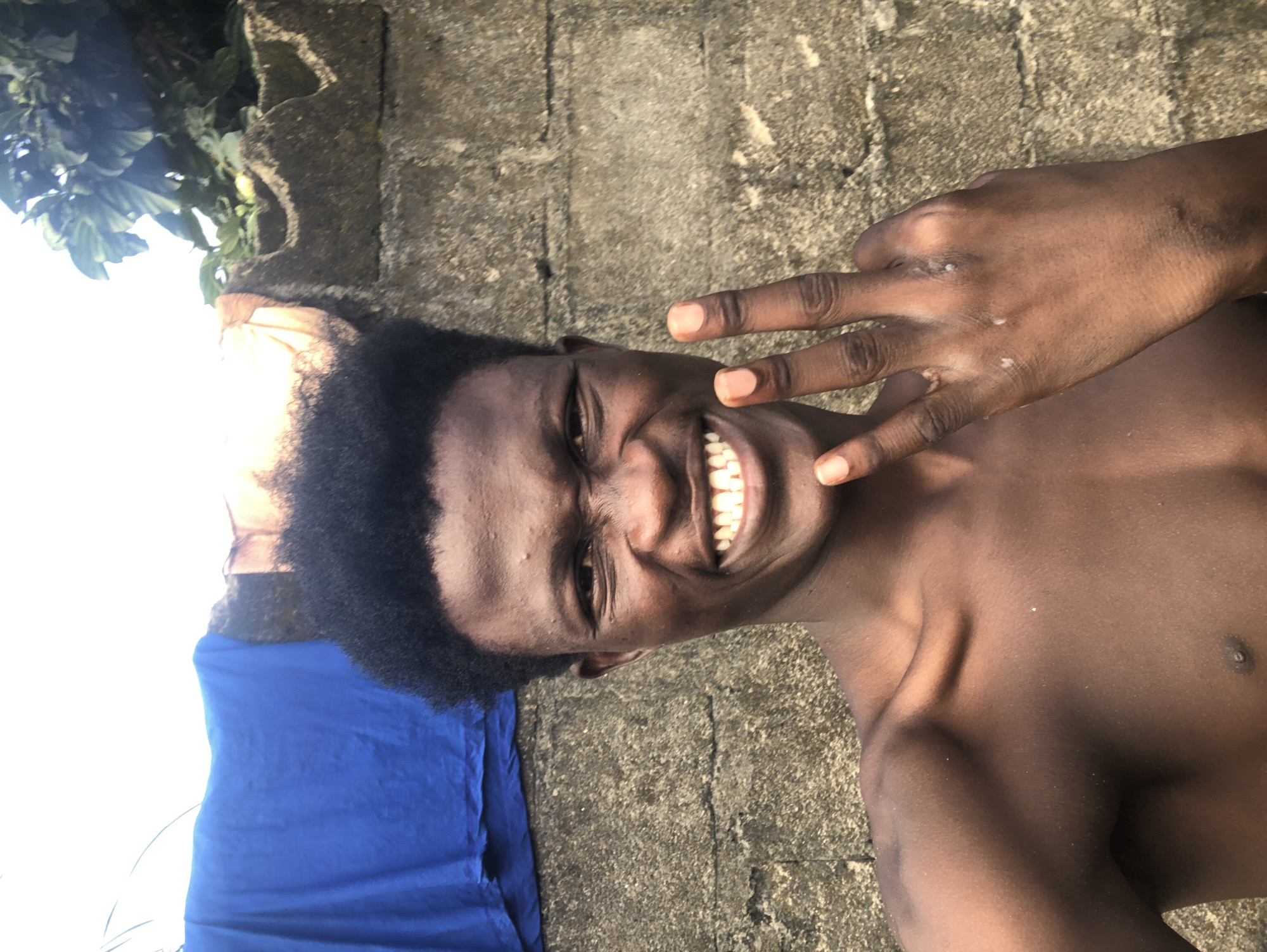 A young man with dark skin and short, black hair smiles broadly at the camera, his teeth bared. He is shirtless, and his left hand is raised, fingers spread in a peace sign. The man is positioned in front of a textured concrete wall. To his left, a bright blue cloth hangs, partially obscuring the wall. Behind him, green foliage and a lighter-colored material are visible above the wall. The lighting suggests an outdoor setting, possibly in Ado, Nigeria, given the overall appearance of the environment and the subject.