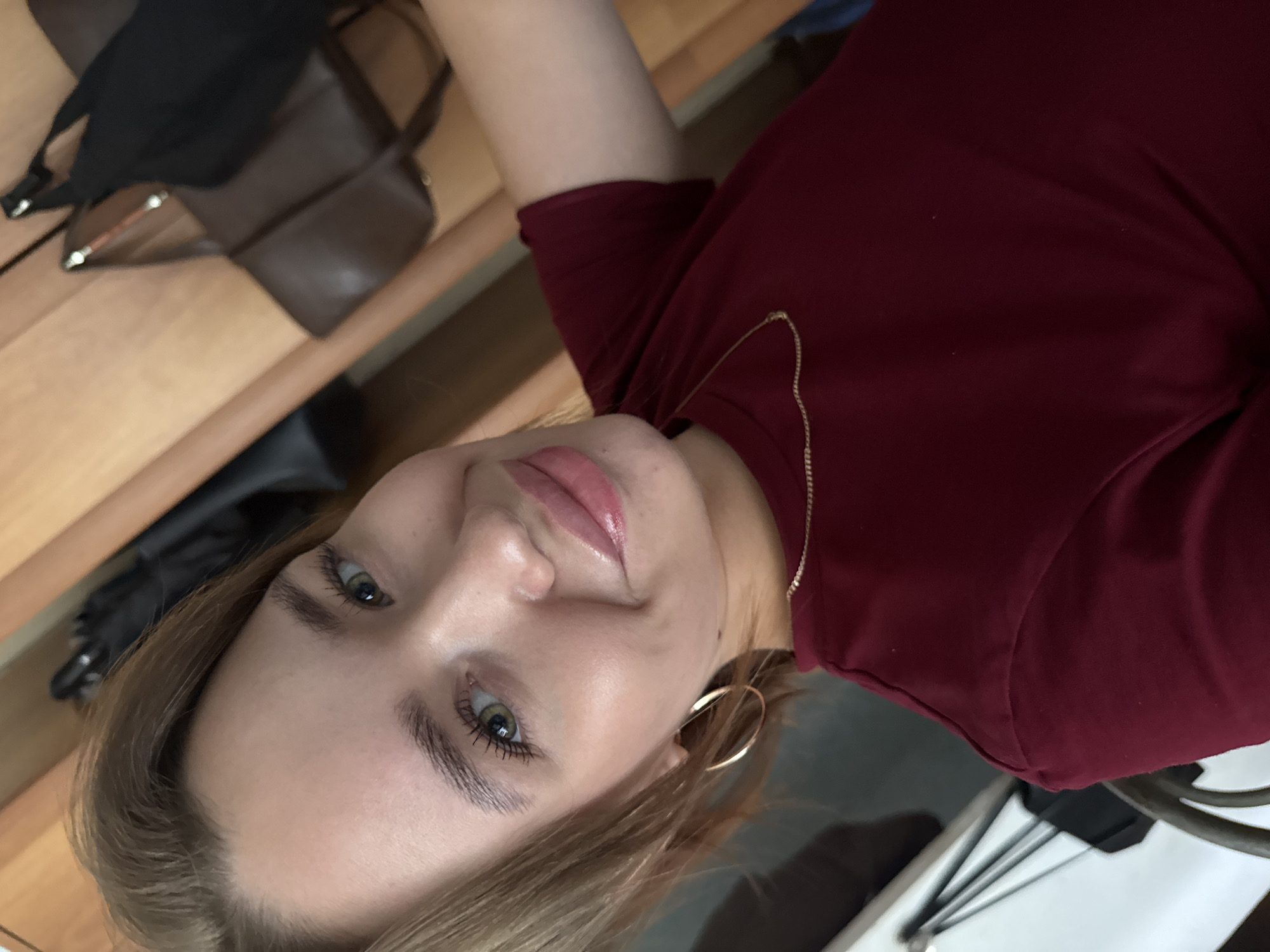 The image shows a young woman with blonde hair, fair skin, and green eyes. She is wearing a maroon t-shirt and a thin gold necklace. She has large hoop earrings. Her lips are glossy. The background appears to be indoors, possibly a closet or a room with wooden furniture and hanging bags. There is no indication of the city of Chaikovsky, Russia, in the image.