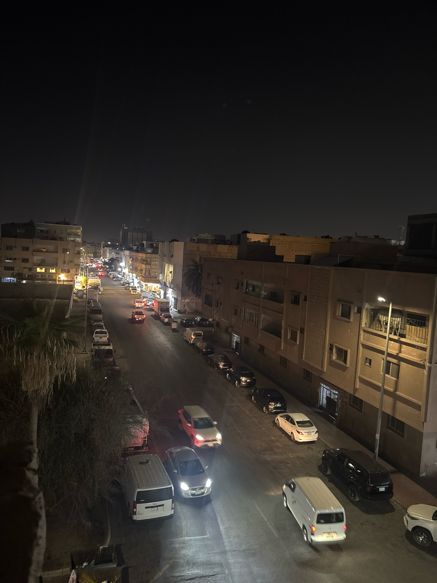 The image depicts a nighttime view of a street in Dammam, Saudi Arabia. The street is lined with multi-story buildings on both sides, characterized by their beige and brown facades, with many windows and balconies visible. Vehicles are parked along the sides of the street and a few are in motion on the road. The lighting suggests it is well past dusk, with streetlights and illuminated building windows casting a warm glow on the scene. The sky is dark, with a faint light beam visible, possibly from a distant light source. The presence of parked cars, buildings, and the general streetscape indicates a residential or mixed-use urban area.