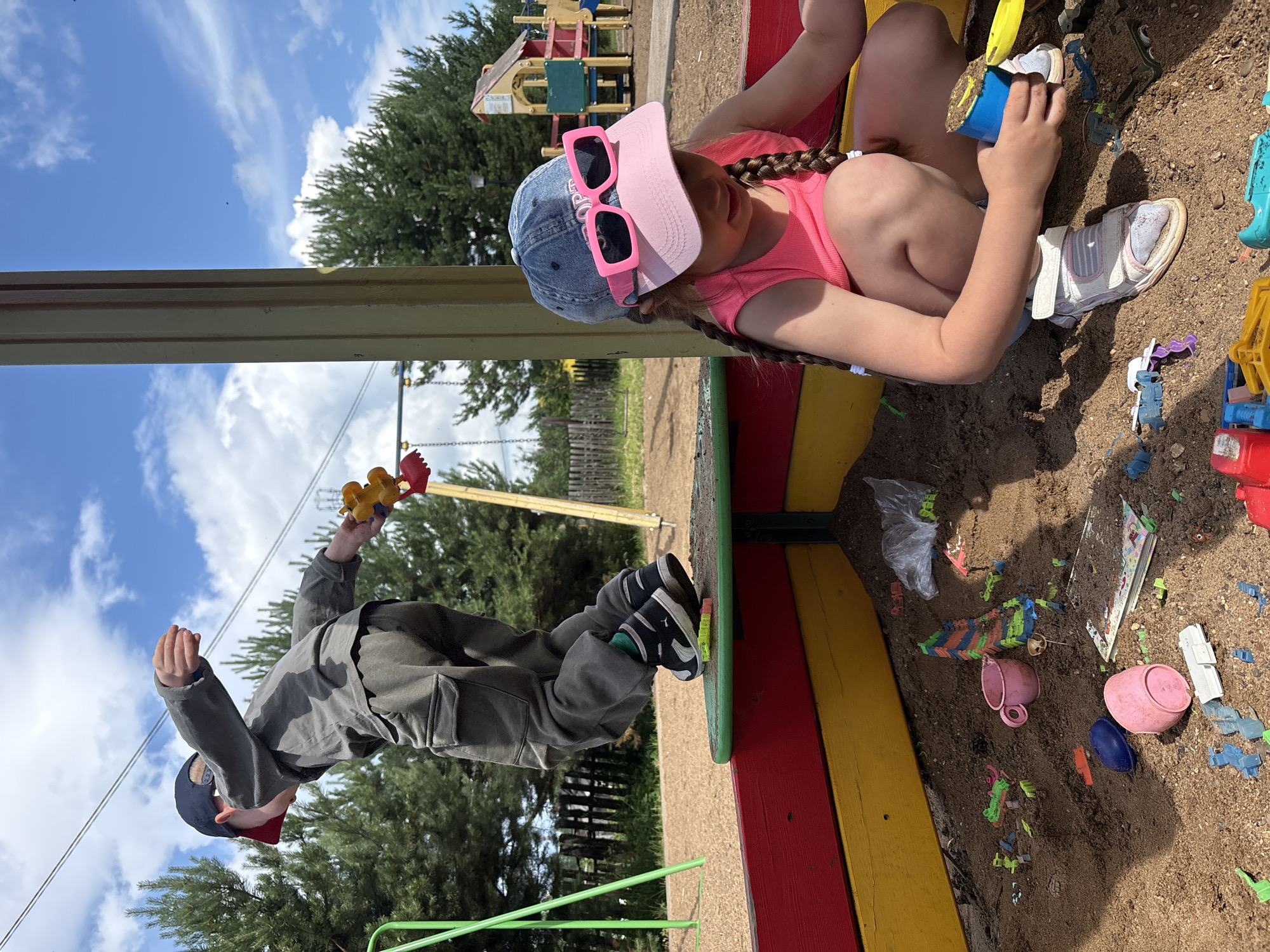 In Chaikovsky, Russia, a boy wearing a gray long-sleeved shirt, gray pants, and a black baseball cap is mid-air, jumping off a green platform that is part of a sandbox. He is holding a yellow toy in his right hand. Below him, a young girl with braided hair, wearing a pink tank top, a denim baseball cap with pink sunglasses, and white sandals, is kneeling in the sandbox, playing with sand. The sandbox has red and yellow painted sides. The ground around the sandbox is covered in sand and woodchips. In the background, there are trees, a wooden fence, and playground equipment, including swings and a play structure with a red roof. The sky is blue with scattered white clouds. The scene captures children engaged in outdoor play at a playground.