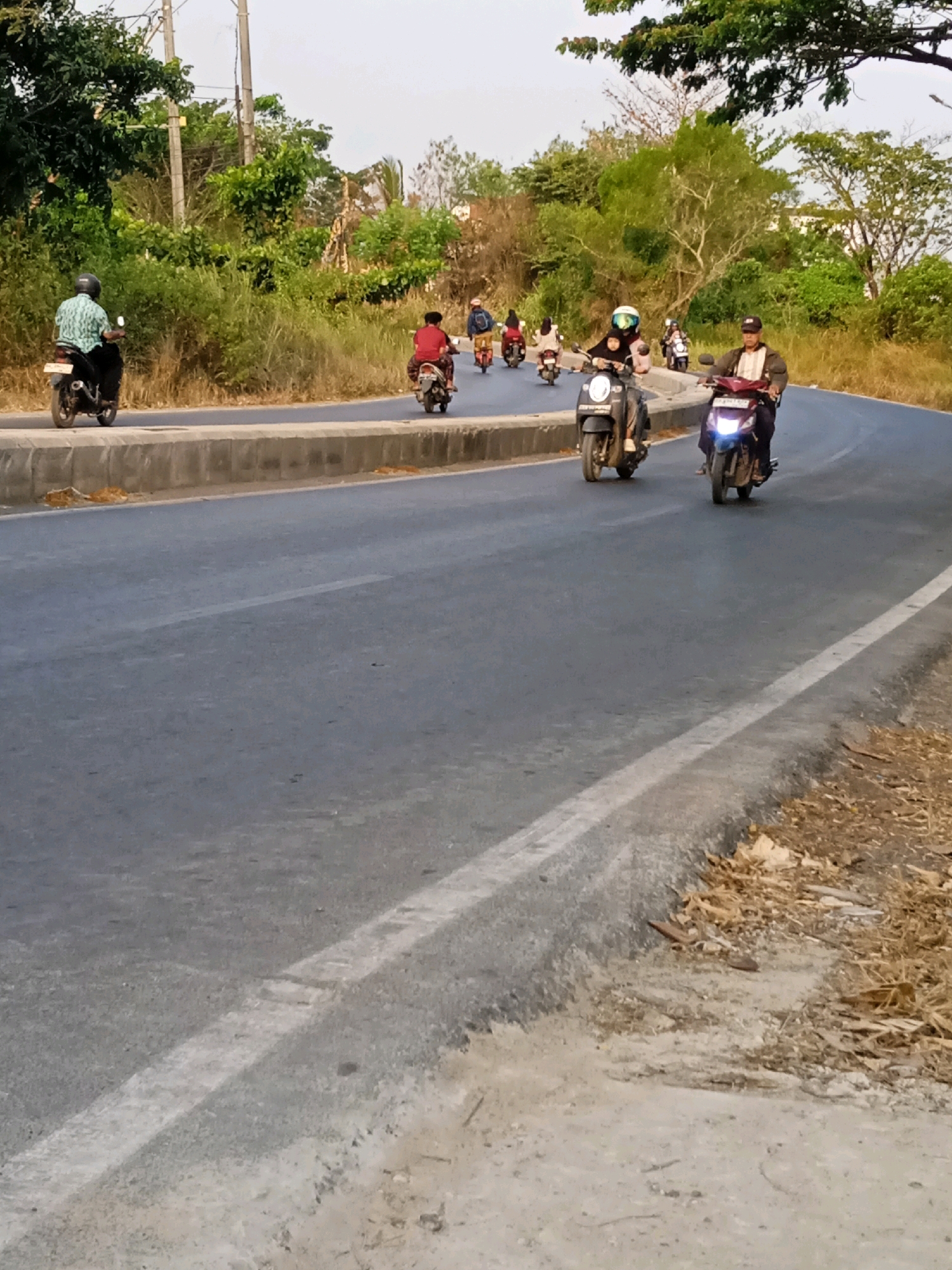 The image depicts a group of people riding motorcycles on a paved road that curves to the right. The road appears to be a two-lane road with a concrete barrier on the left side. The surrounding environment is characterized by lush green vegetation on the left and dry, brown grass and trees on the right. The sky is overcast, suggesting it might be late afternoon or early morning.
In the foreground, the asphalt road is visible with a white lane marking. The concrete barrier separating the road from the embankment is also clearly visible.
Several individuals are riding motorcycles in various positions on the road. On the far left, a person in a light blue and green patterned shirt and black pants is riding a black motorcycle. Further down the road, a person in a red shirt is riding a light-colored motorcycle. To the right of them, a person in a blue jacket and black pants is riding a red motorcycle. In the middle of the frame, a person wearing a helmet with a white visor and a dark jacket is riding a black scooter, with a passenger behind them. To the right of this scooter, a person in a light-colored shirt and dark pants is riding a red motorcycle with its headlights on. Further in the distance, more motorcycles and riders can be seen, some blurred by motion.
The overall scene suggests a typical day of transportation or leisure activity in a semi-urban or rural setting in Banjarmasin, Indonesia, where motorcycles are a common mode of transport. The presence of trees, vegetation, and the style of the road and motorcycles are consistent with locations in that region.