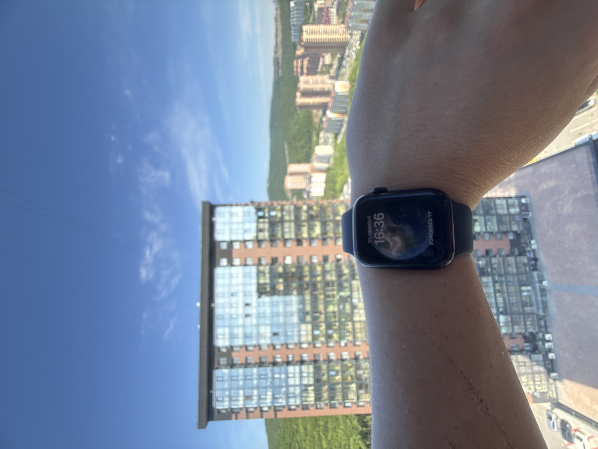 The image shows a close-up of a person's arm wearing a smartwatch. The smartwatch displays the time as 16:36 and indicates "POCO 19°". The background is out of focus, but it appears to be a city scene with tall buildings, greenery, and a clear blue sky, suggesting it is daytime and the weather is fair. A scar is visible on the person's forearm. The orientation of the image is rotated, with the sky and buildings appearing to the left and right of the arm. The context provided suggests the location is Vladivostok, Russia. No specific activity or event is evident, other than the person wearing a smartwatch while observing the surroundings.
