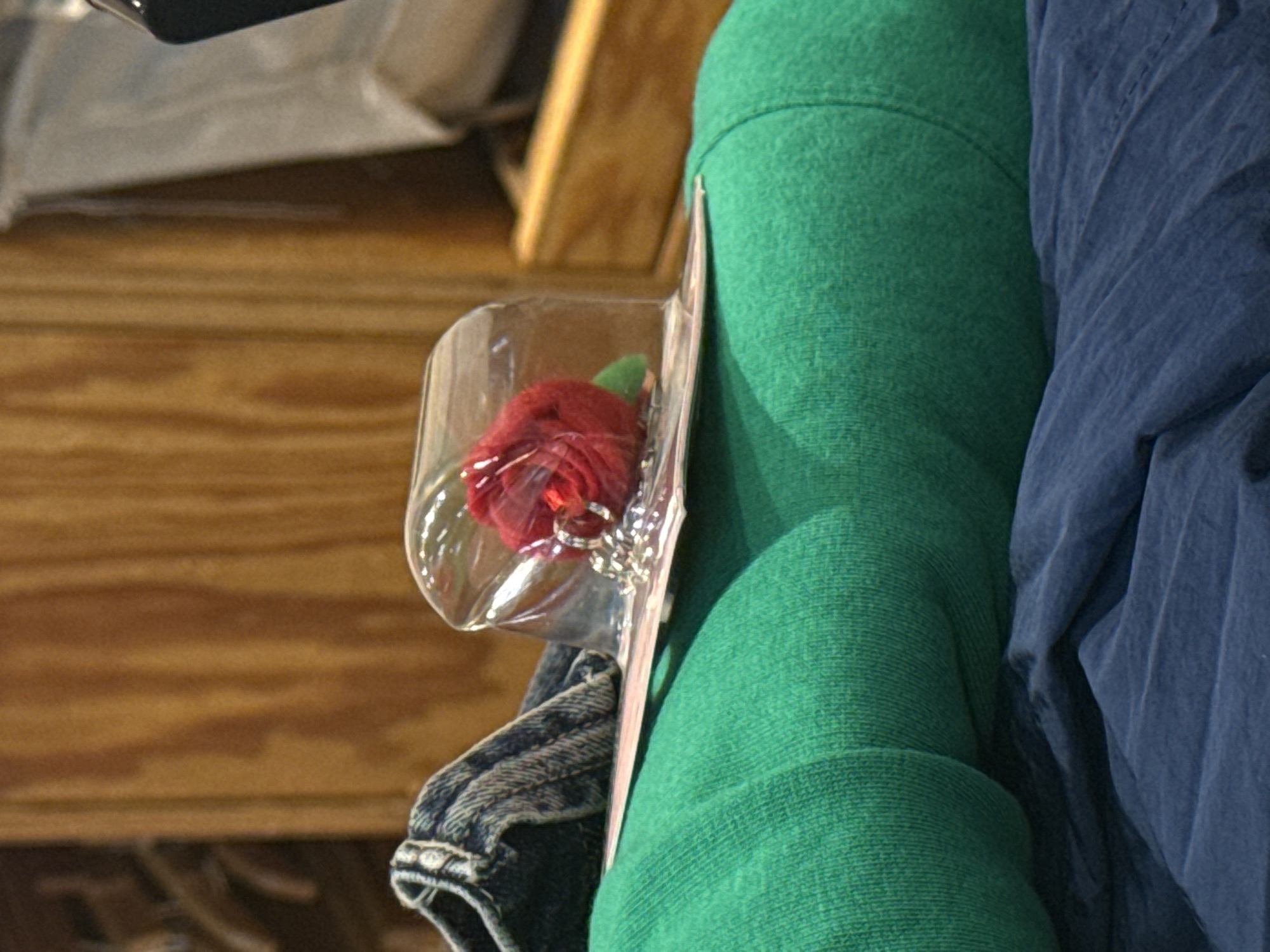 A still life shows a packaged red rose with a silver necklace lying against folded clothes. The rose is encased in a clear plastic bubble and mounted on a pink card. The clothing includes a green sweater, a blue shirt, and a piece of denim. The background shows a wooden surface. The location is not discernible from the image.