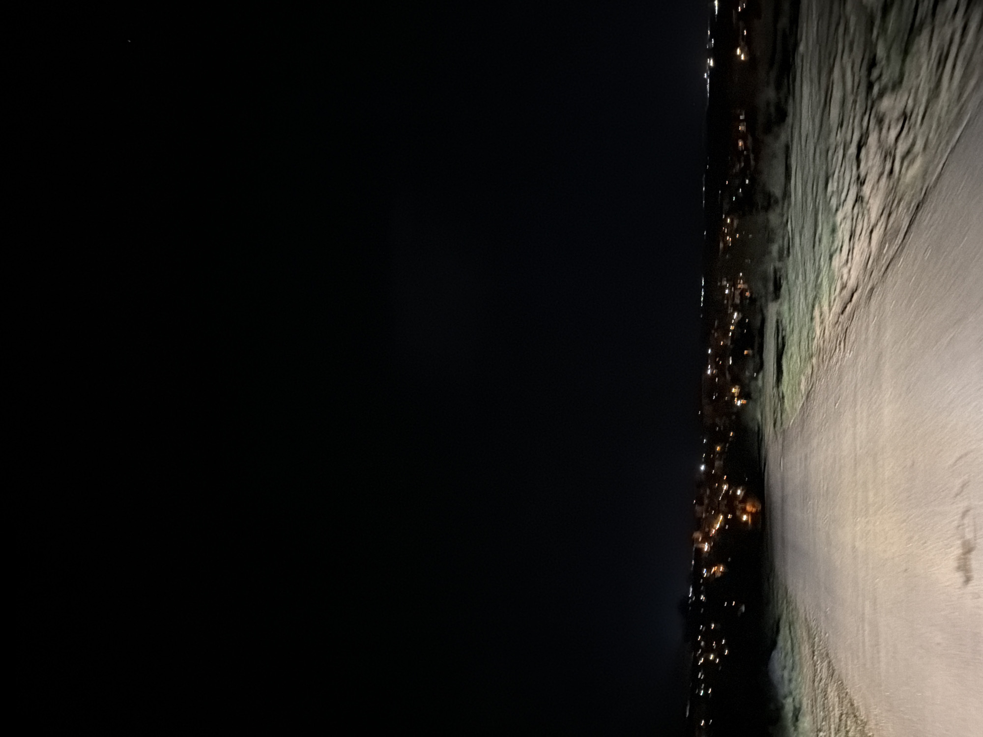 The image depicts a nighttime scene. On the right, a road or paved surface is visible, illuminated by a light source, likely headlights. To the left of the road appears to be a grassy or natural terrain, possibly a hillside. In the distance, a series of lights, suggesting a city or town, are scattered along the horizon. The sky is dark.
Given the mention of Sofia, Bulgaria, and the presence of an elevated road or path overlooking a city, this could potentially be a road leading to or from the city, offering a panoramic view. However, without further details, pinpointing the exact location is not feasible.