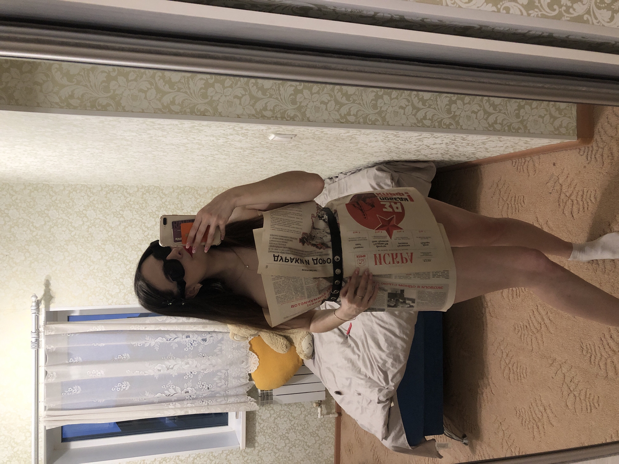 A mirror selfie captures a young woman posing creatively in what appears to be a bedroom. She has long, dark hair and wears dark sunglasses. With her right hand, she holds a smartphone to take the picture, while her left hand helps secure several sheets of newspaper wrapped around her torso, appearing to form a temporary outfit or prop. A black belt with metal studs is visible over the newspaper. She wears white socks on her feet, and her legs are visible.
The background shows a domestic setting. Behind the woman, there's a bed with white bedding and a blue base, on which a yellow plush toy rests. To the left, a window with white, patterned curtains is visible, along with a white radiator below it. The walls are covered in two different patterns of wallpaper – a lighter, intricate floral design above a simpler textured pattern. The floor is carpeted in a light brown, textured pattern.
The visible text on the newspaper pages is in Russian and includes words such as "МОСКВА" (Moscow), "УДЕРЖАН" (Detained), "РАЗБОЙ" (Robbery), "КУБАНЬ" (Kuban), "ПОБЕДА" (Victory), "ГАЗЕТА" (Newspaper), and "АЗБУКА" (Alphabet). A red star emblem is also prominent on one section of the newspaper. The smartphone case also shows partial text, "INS". The time of day is not explicitly clear. The woman's pose and creative use of newspaper imply a playful or artistic mood in this indoor setting, located in Noviy, Russia.
