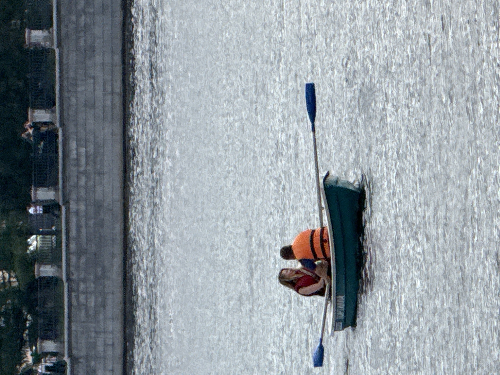 The image shows two people in a rowboat on a body of water. The person in the front is wearing a red life jacket and the person in the back is wearing an orange life jacket. They are both facing each other and appear to be talking. There is a paddle in the water to the left of the boat. In the background, there is a stone wall and trees. The overall scene suggests a leisurely activity on a waterway. The location appears to be a city park or waterfront area in Yekaterinburg, Russia, possibly near the Iset River or one of its surrounding water bodies.