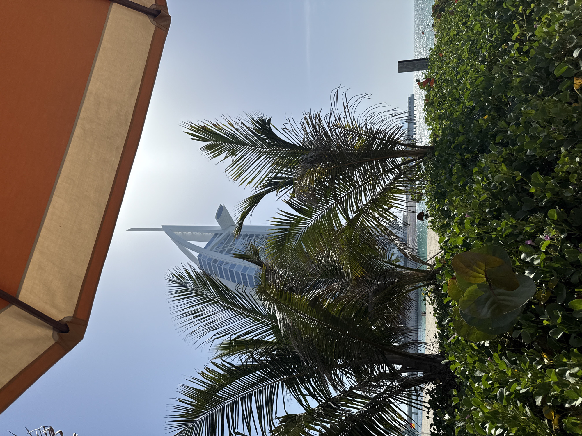 The image captures a daytime scene in Dubai, United Arab Emirates, where the Burj Al Arab hotel is visible through palm trees. The view is partially obstructed by an orange and beige-striped sunshade on the left side of the frame. Lush green foliage fills the right side. The sky is bright and clear. The beach and ocean are visible in the background. A dock structure is apparent in the distance.