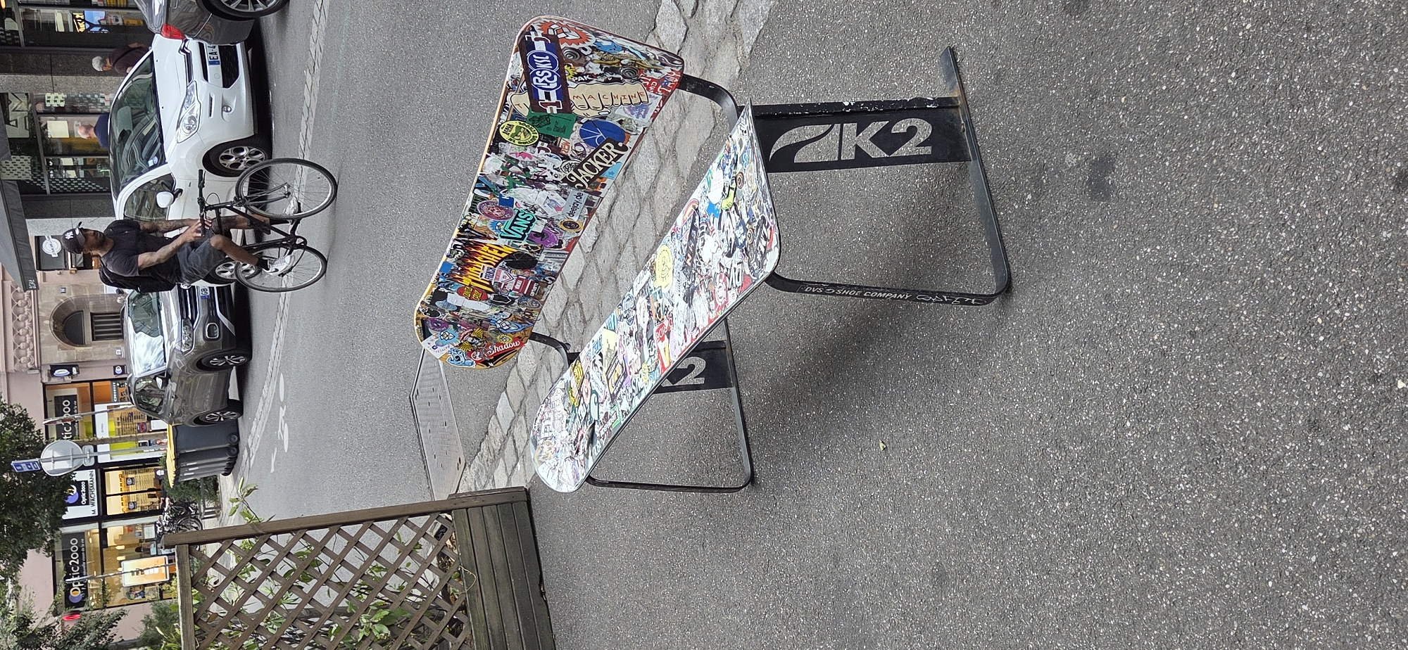 This is a street-level, eye-level, medium shot of a bench designed to resemble snowboards, covered in stickers, parked on the sidewalk of a street in Strasbourg, France. A man on a bicycle with a backpack is riding past the bench on the asphalt street. In the background, a gray SUV and a white car are parked, with commercial buildings and trees lining the street. The bench itself is made of black metal legs and snowboard tops as seats, with numerous stickers depicting various brands, logos, and graphics. The street is paved with asphalt, and the sidewalk is made of cobblestones. The lighting suggests it is daytime.