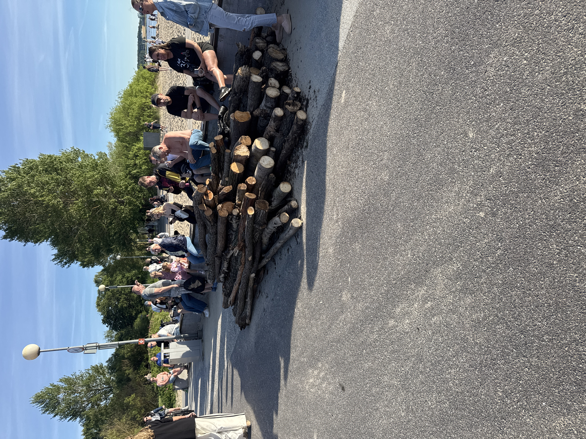In Tallinn, Estonia, a group of people are gathered on a sandy beach area adjacent to a paved walkway. A large pile of chopped wood lies on the ground between the walkway and the beach. Several individuals are seated on a low, concrete wall facing the beach, with some looking towards the water or engaged in conversation. Others are standing or walking on the paved path. The scene is illuminated by the late afternoon sun, casting long shadows. The background features trees, a lamppost, and a clear blue sky. The overall atmosphere appears relaxed, with people enjoying the outdoor space.