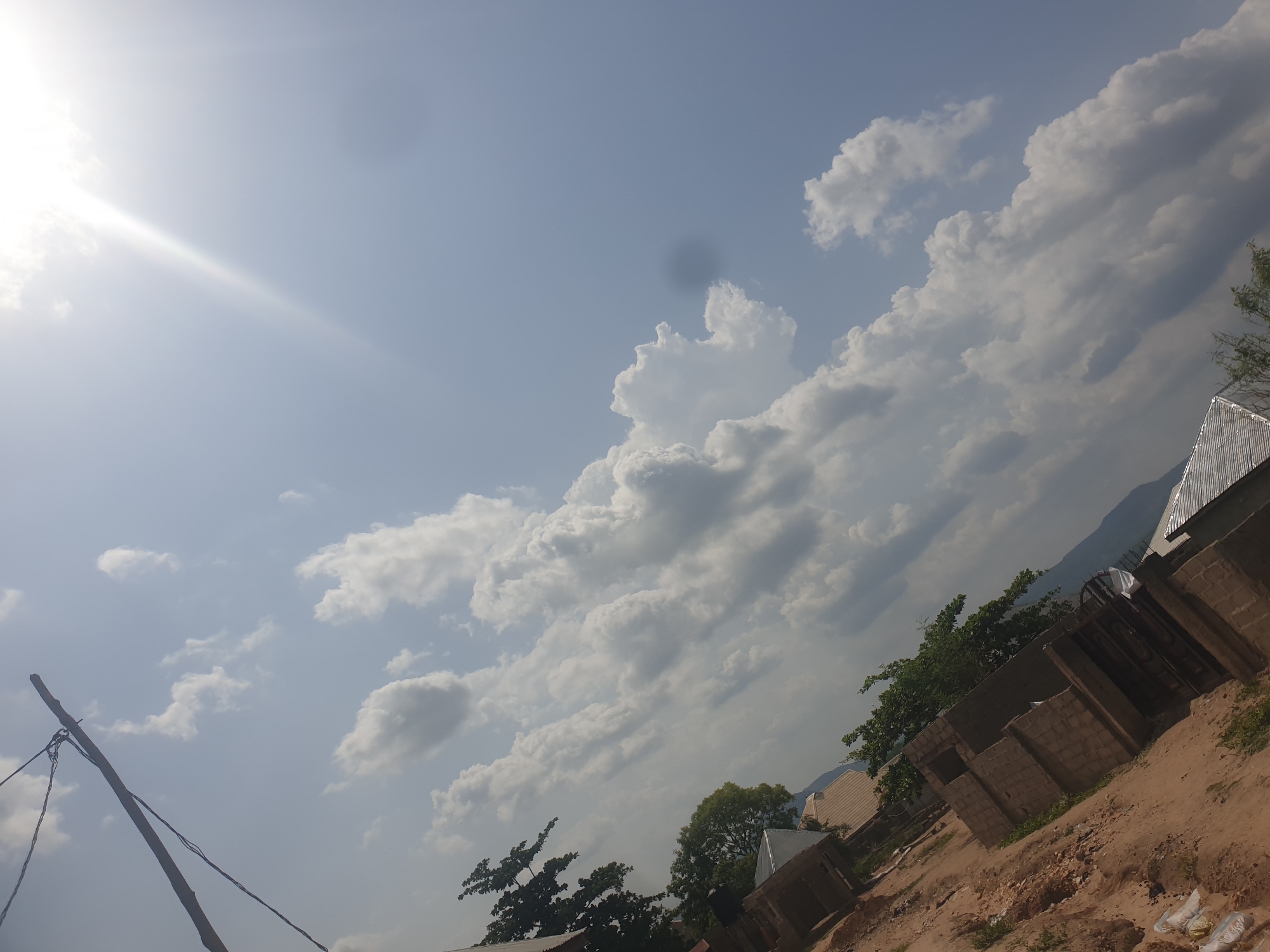 The image shows a partly cloudy sky over Zango-ndaji, Nigeria. In the foreground, there is a dirt ground, with the start of the construction of houses. Also in the foreground are trees. The sky is blue with bright sunlight.