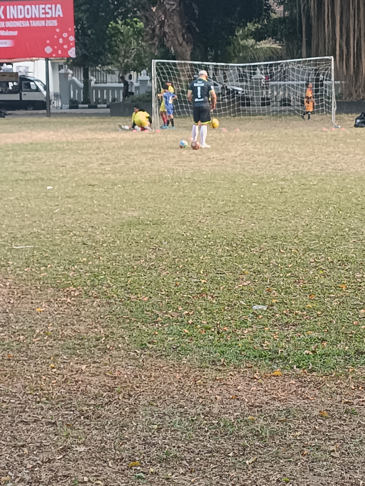 In Sleman, Indonesia, on a grass field, a group of young soccer players practices near a goal. One player in a black and gray jersey with the number "1" on the back stands facing away from the camera, with a soccer ball at his feet. Another player in a yellow jersey is on the ground near the goal. A player in a blue jersey is near the goalpost. A player in an orange jersey is in the goal. The field has several orange cones placed on it. In the background, a red banner with white lettering is visible. The scene appears to be a soccer training session.
