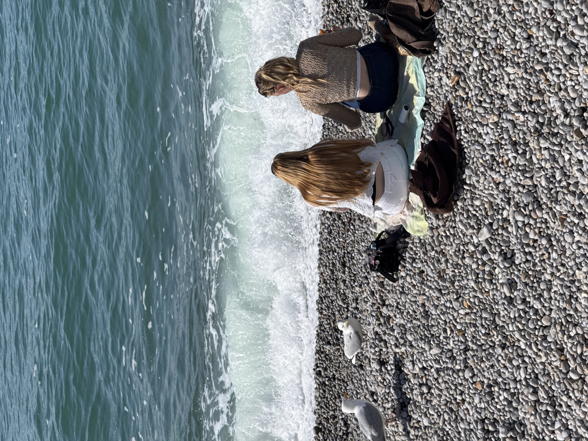 The image captures a sunny scene on the pebble beach of Étretat, France. Two women are observed from behind, seated on a light green and yellow striped blanket, facing the ocean. The woman on the left has long, straight, reddish-blonde hair and wears a white long-sleeved top and white shorts. The woman on the right has blonde hair tied in a ponytail and wears a light brown crocheted long-sleeved top exposing her lower back, paired with dark blue jeans; she holds a light-colored object in her right hand.
In the foreground and extending towards the water, the beach is densely covered with smooth, grey and light brown pebbles. To the left of the women, two seagulls stand on the pebbles; the closer one faces slightly right, and the further one faces slightly left. A black handbag is positioned on the pebbles behind the right woman, and a dark brown garment is folded on the blanket in front of her. Another dark bag is visible on the far right.
The ocean displays a deep blue-green hue, with a significant wave breaking directly in front of the women, creating a prominent line of white foam and spray across the middle ground. The water exhibits surface ripples and scattered patches of foam.