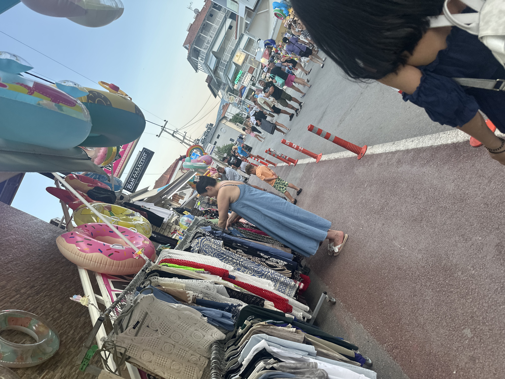 The image captures a lively street scene in Leptokarya, Greece, on a clear day. A woman wearing a long blue dress is browsing clothes displayed on a rack outside a shop. She is examining a white lace top. The street is lined with shops and stalls, displaying a variety of items including inflatable rings designed to look like donuts and other shapes. People are walking along the street, some appear to be tourists. In the background, buildings with balconies are visible, and power lines run overhead. The ground is paved with red asphalt.