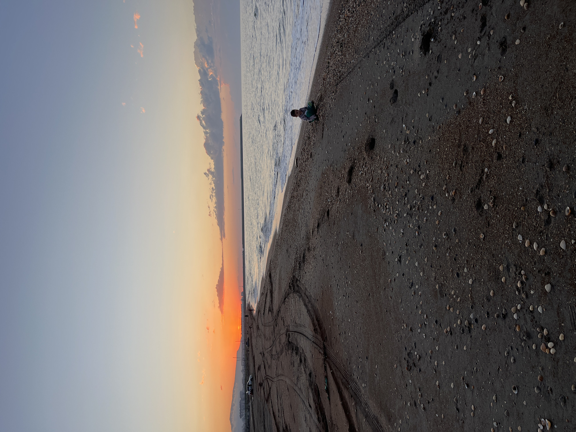 A medium-long outdoor shot reveals a beach at sunset in Kaspiysk, Russia. The sky transitions from blue to orange and red with clouds on the left, indicating the setting sun. The sea occupies the central portion of the image, with waves breaking on the shore. On the beach, tire tracks are visible in the dark sand, which is also scattered with shells and pebbles. In the upper right area of the shot, a child is kneeling on the sand at the shoreline, facing the viewer. A light-colored vehicle and part of a fence can be seen to the lower left. Mountains rise in the distance at the bottom of the image.