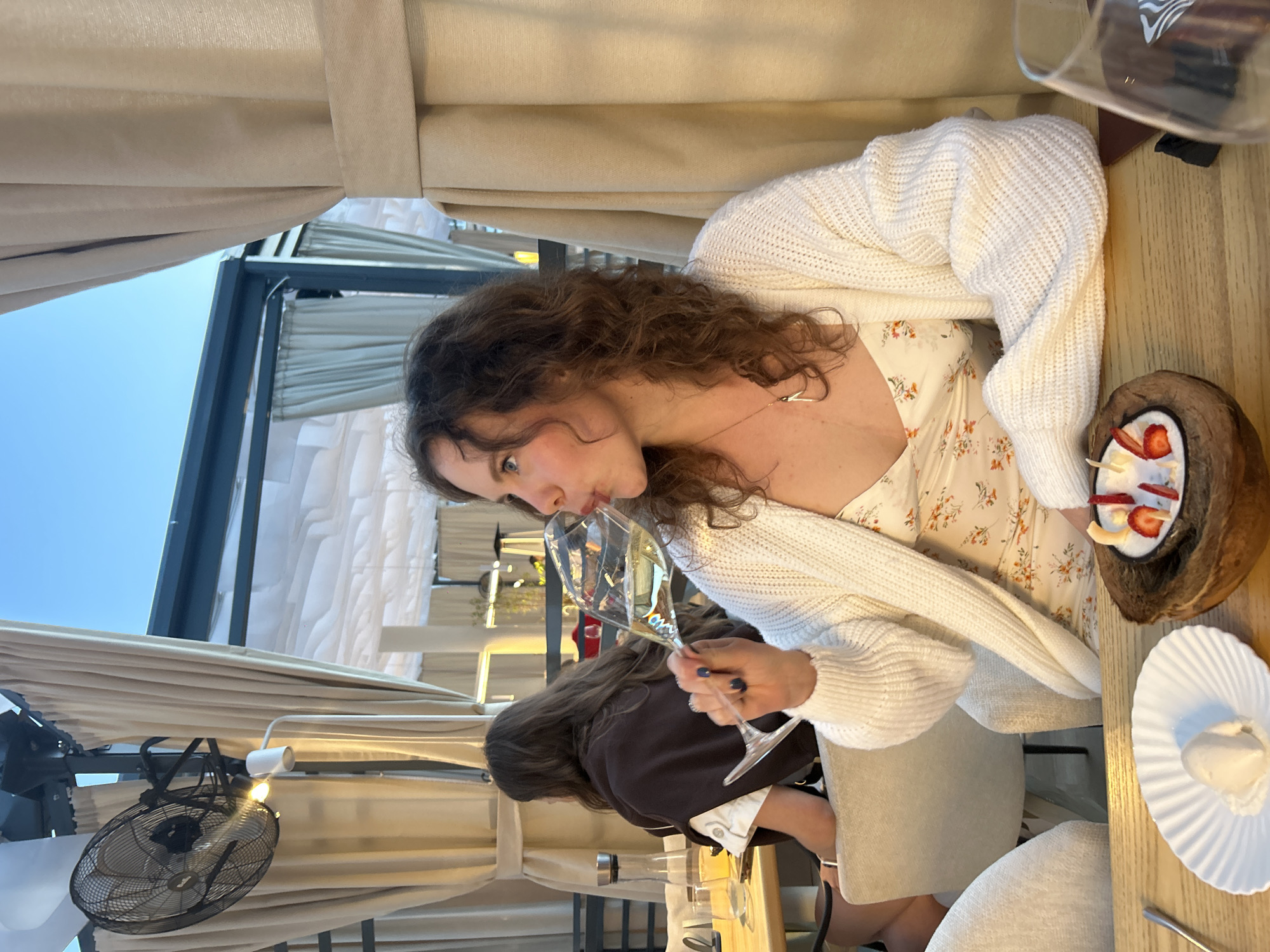 The image depicts a woman enjoying a drink at an outdoor or semi-outdoor dining establishment, likely a restaurant patio, in Krasnoyarsk, Russia. The main subject, a woman with curly brown hair, is seen in profile, wearing a white floral dress and a cream-colored knitted cardigan. She is holding a champagne flute and taking a sip of a light-colored sparkling beverage, her expression appearing relaxed and focused on her drink.
The setting includes light-colored retractable curtains or blinds, suggesting a covered yet open-air space. A clear blue sky is visible in the background, indicating fair weather, possibly late afternoon or early evening. On the wooden table in front of her are desserts: one served in a natural coconut shell, adorned with white cream, sliced strawberries, and coconut flakes, and another scoop of ice cream or sorbet on a white fluted plate with a spoon.
Another person with dark hair is partially visible seated across the table. Other elements in the scene include a black oscillating fan attached to the patio structure and a modern light fixture. The overall atmosphere is one of leisure and enjoyment. No visible text is present in the image.